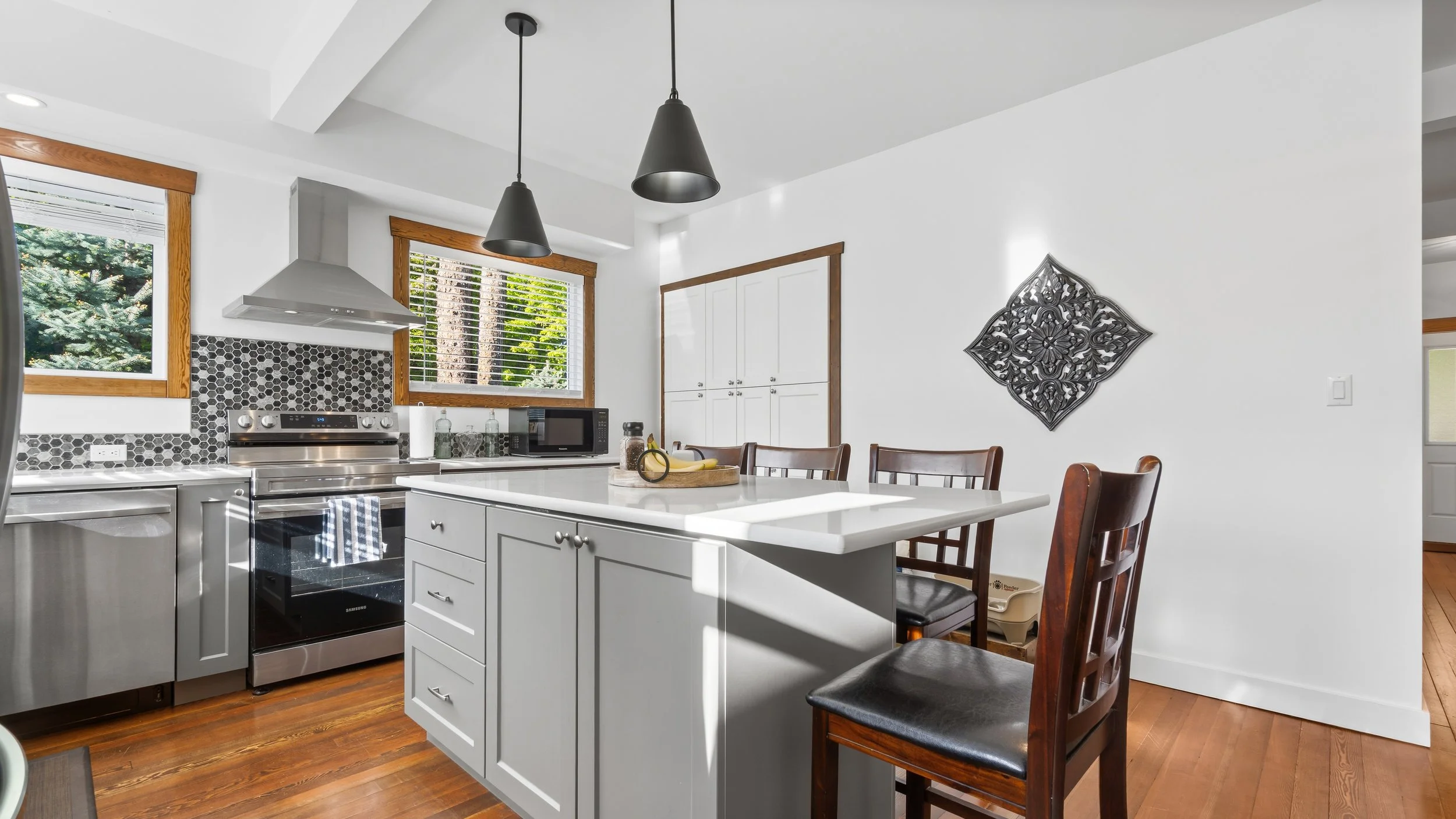 A modern kitchen with white cabinets, a white countertop, two windows with wooden trim, stainless steel appliances, black and white patterned backsplash, black pendant lights, and a dining table with wooden chairs.