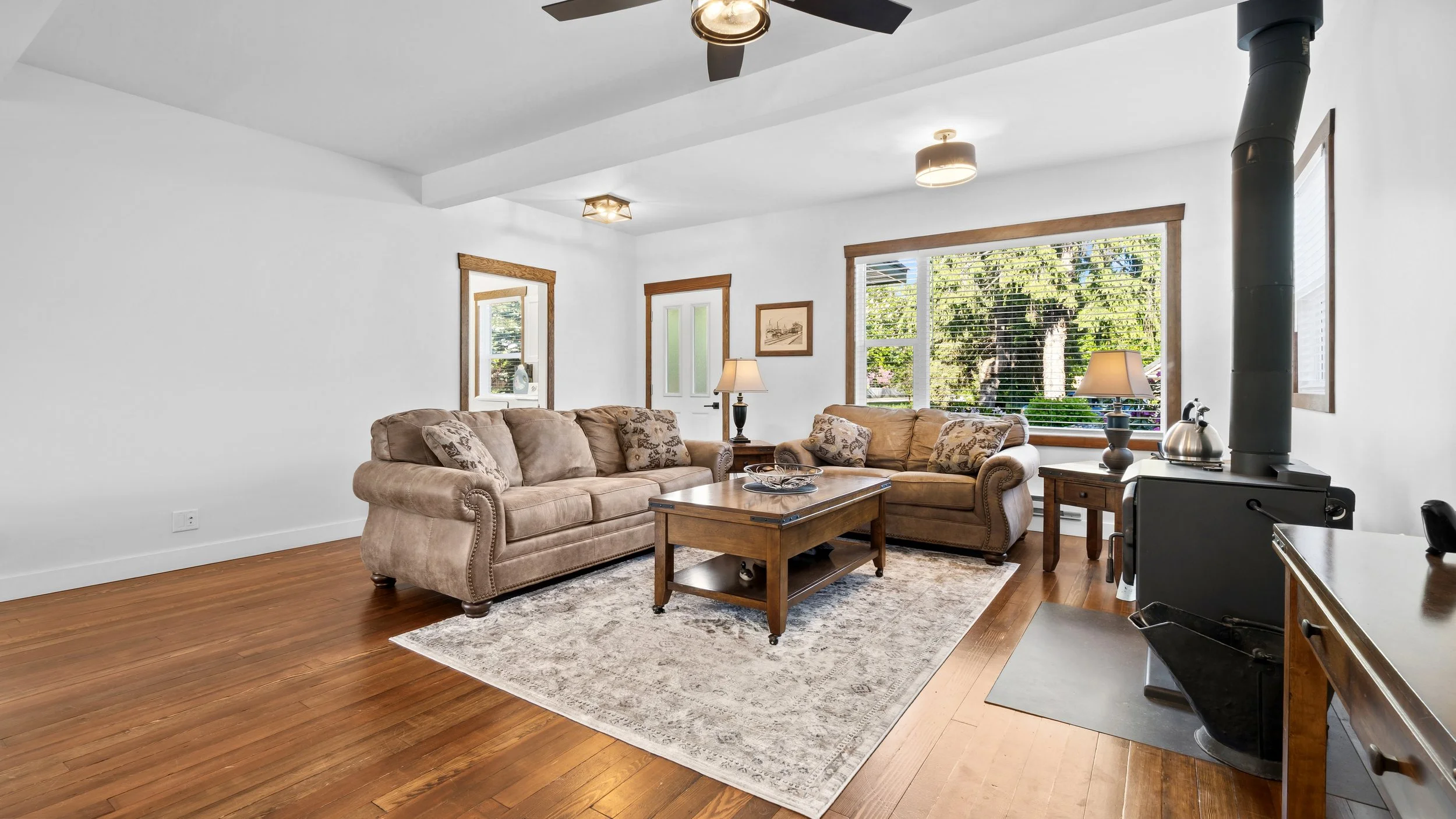 Living room with two beige sofas, a wooden coffee table, a side table with a lamp, large window with blinds, and wood flooring.