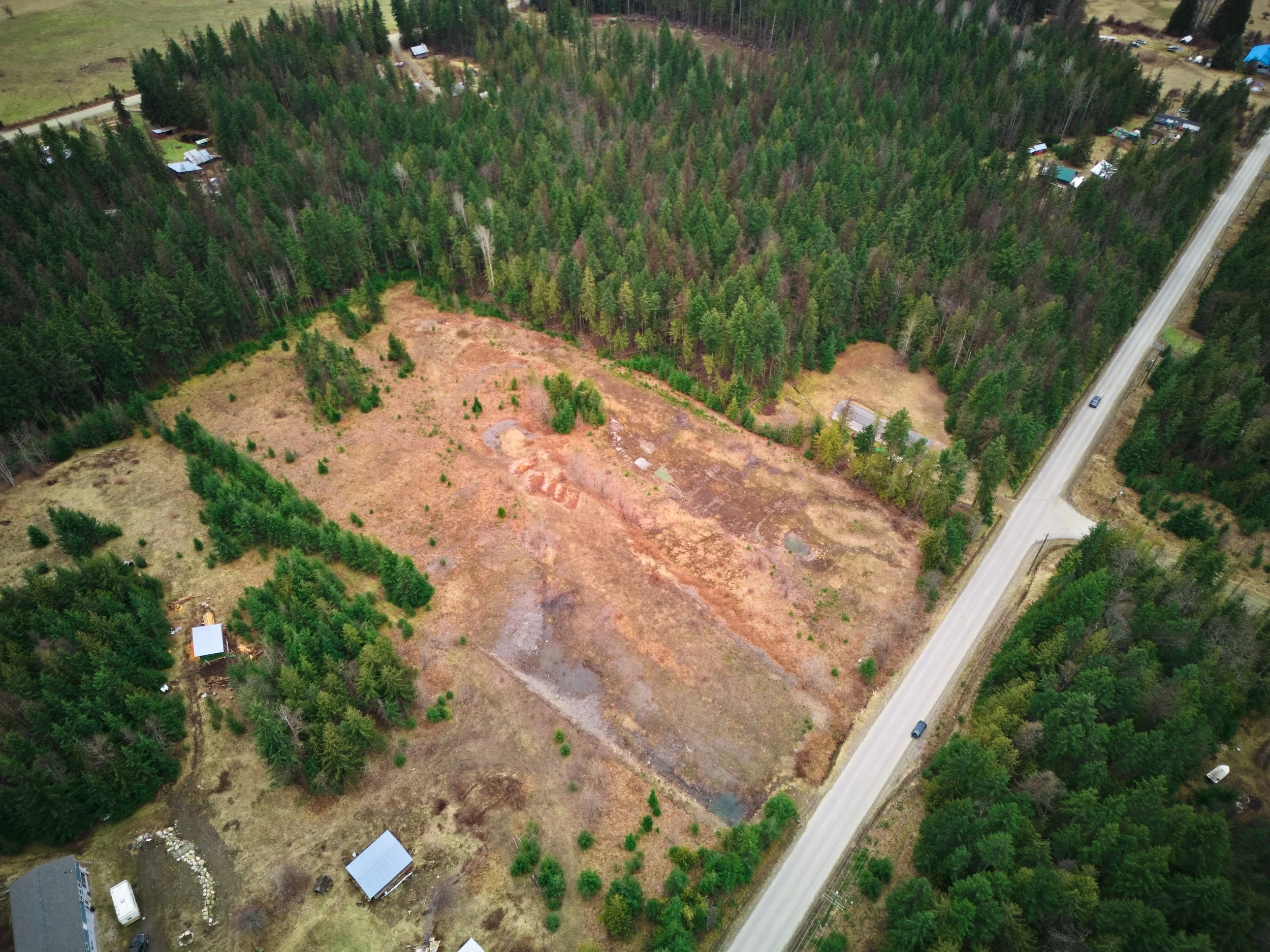 Aerial view of a partially cleared plot of land surrounded by dense forest, with a road running along one side and a few small buildings nearby. Nakusp Real Estate Photography