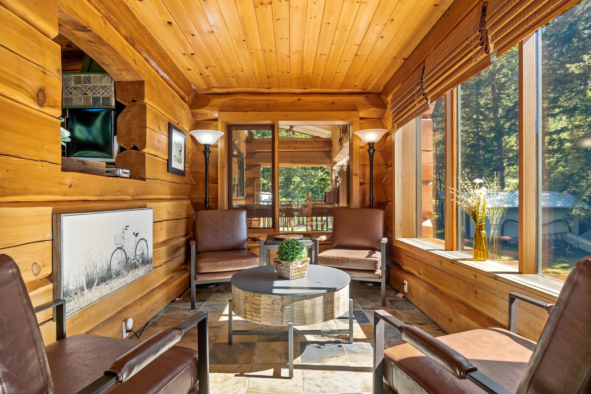 Interior of a cozy wooden cabin room with large windows, featuring leather chairs, a small round table with a plant, and wall art, surrounded by a forest view.