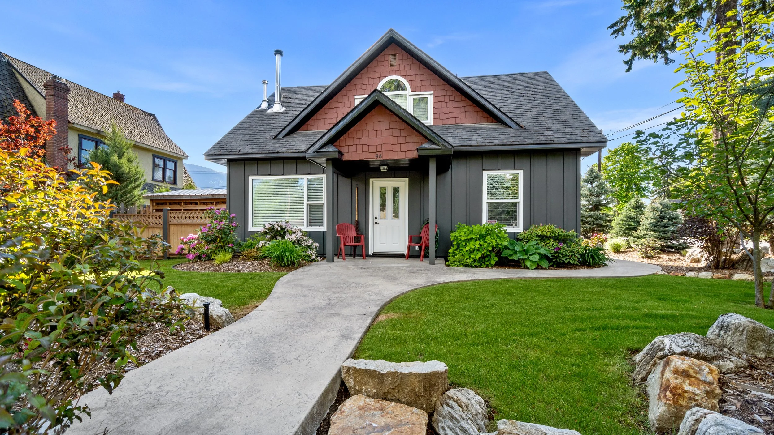 A modern house with a dark gray exterior, a front porch with red chairs, surrounded by a well-maintained green lawn, colorful bushes, and trees under a clear blue sky. Nakusp, 98 2nd Ave
