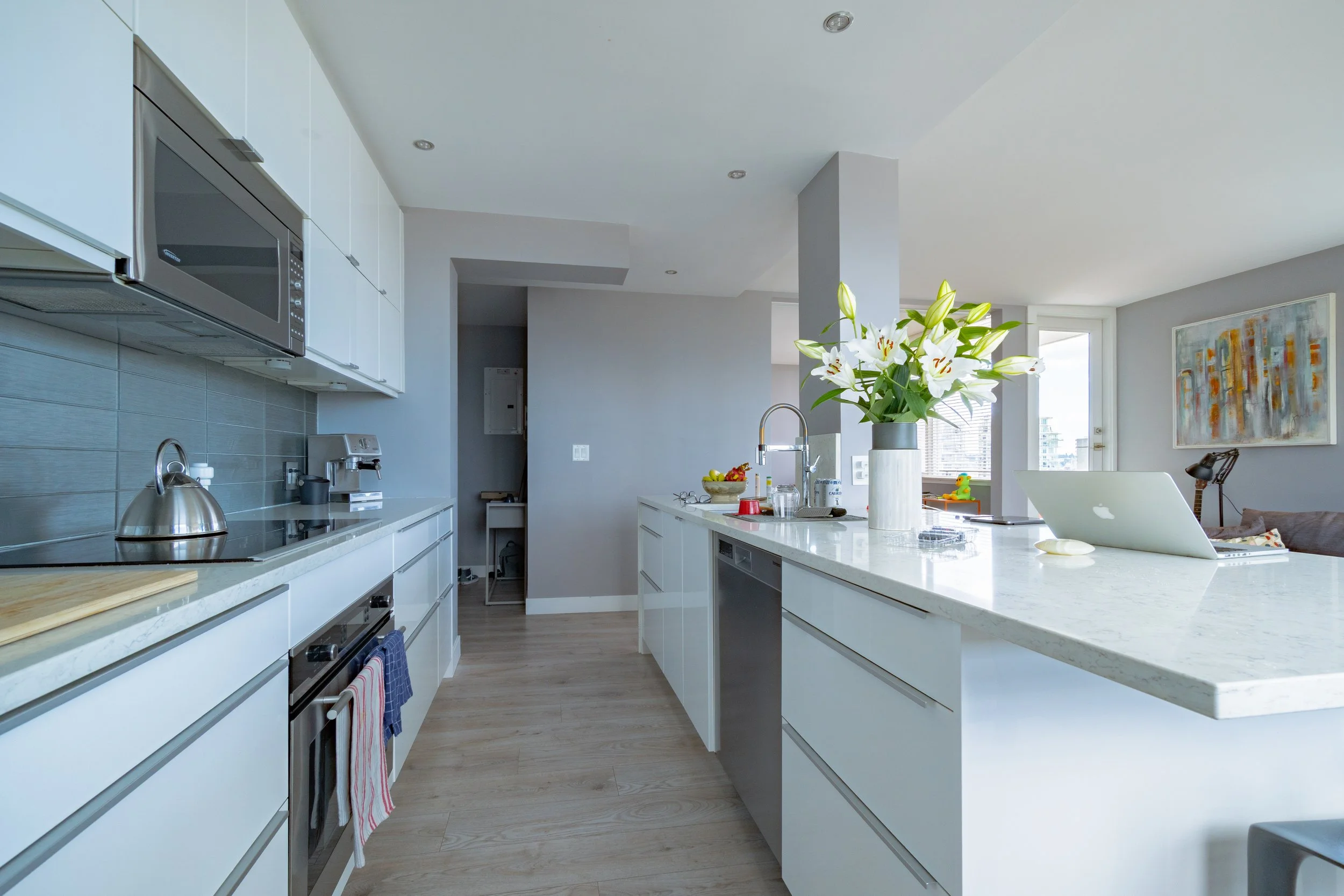 Modern kitchen with white cabinets and a marble countertop, white flowers in a vase, and a laptop on the counter. - Vancouver BC, 1903 Comox St.