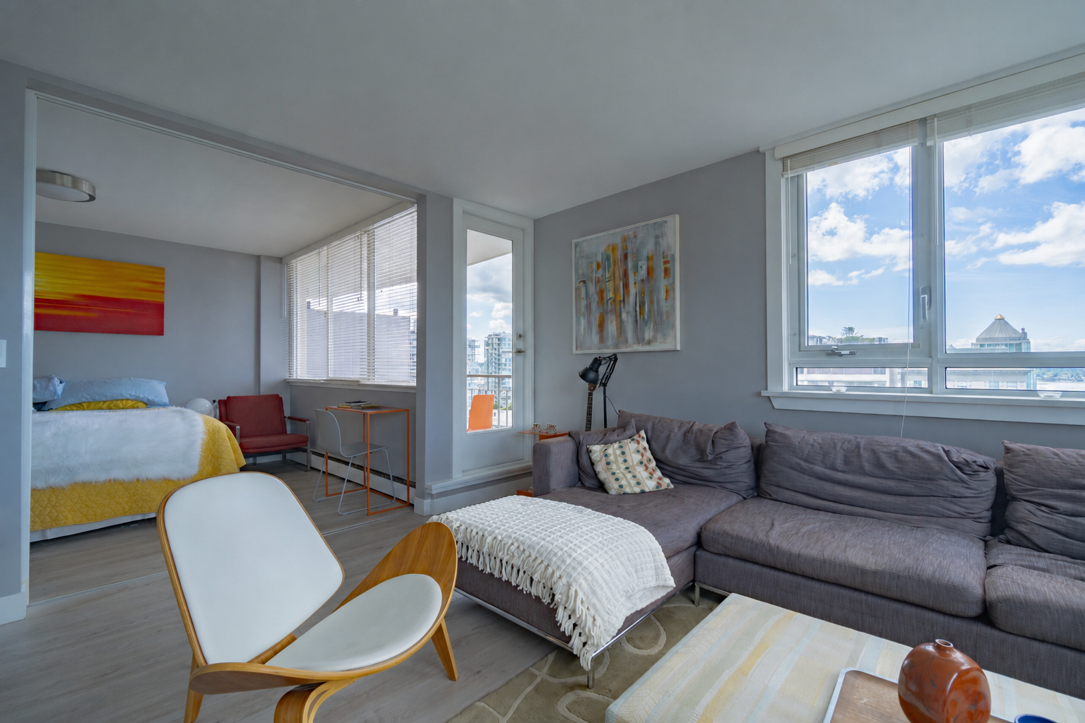 An open-concept living space with a gray sectional sofa, a wooden lounge chair, large windows showing a cityscape, a bedroom with a bed, colorful artwork, and modern decor. - Vancouver BC, 1903 Comox St.