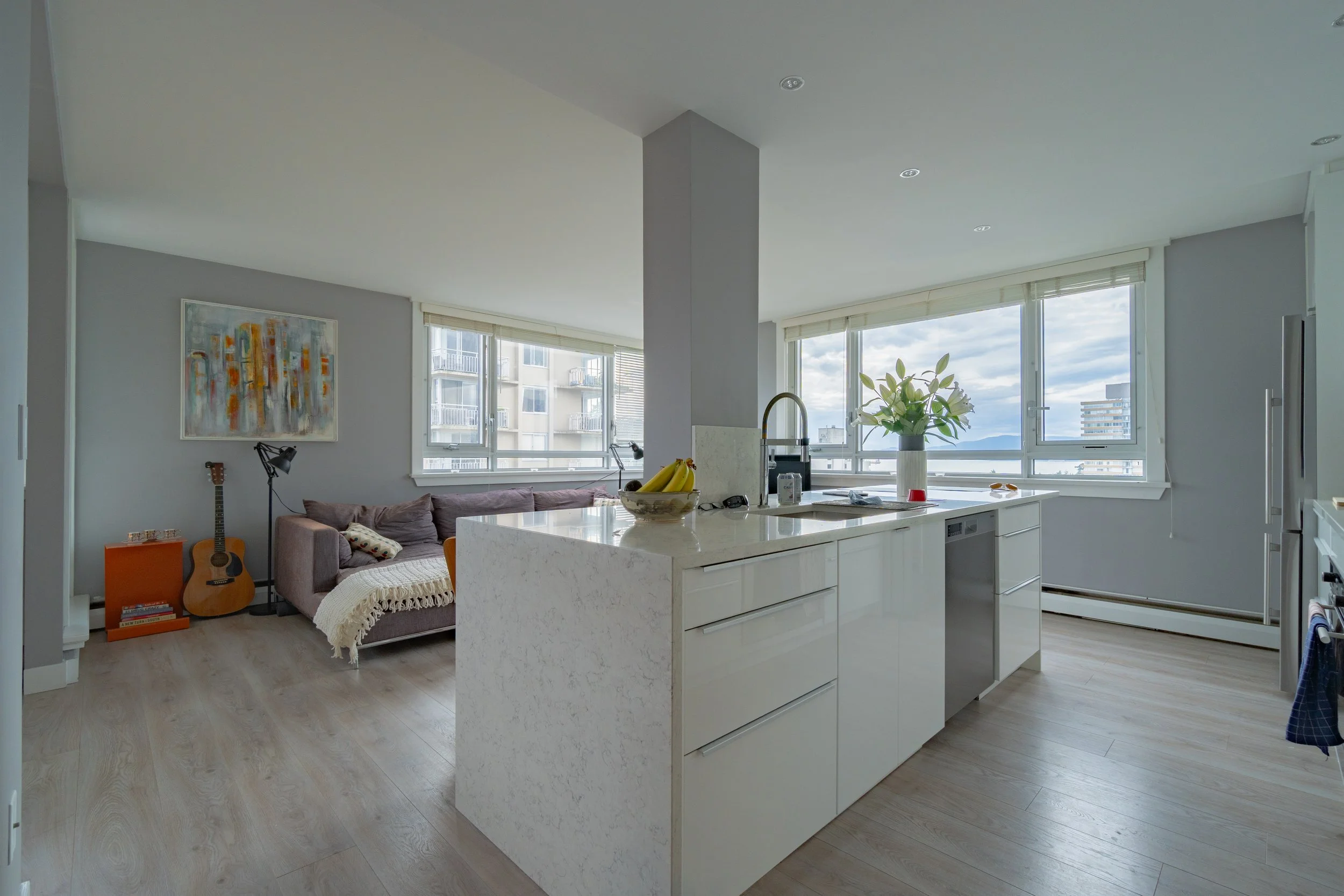 Open-concept apartment with kitchen and living area. Features white cabinetry, a marble island with a sink, large windows providing natural light. Living area has a sofa, a guitar, and artwork on the wall. - Vancouver BC, 1903 Comox St.