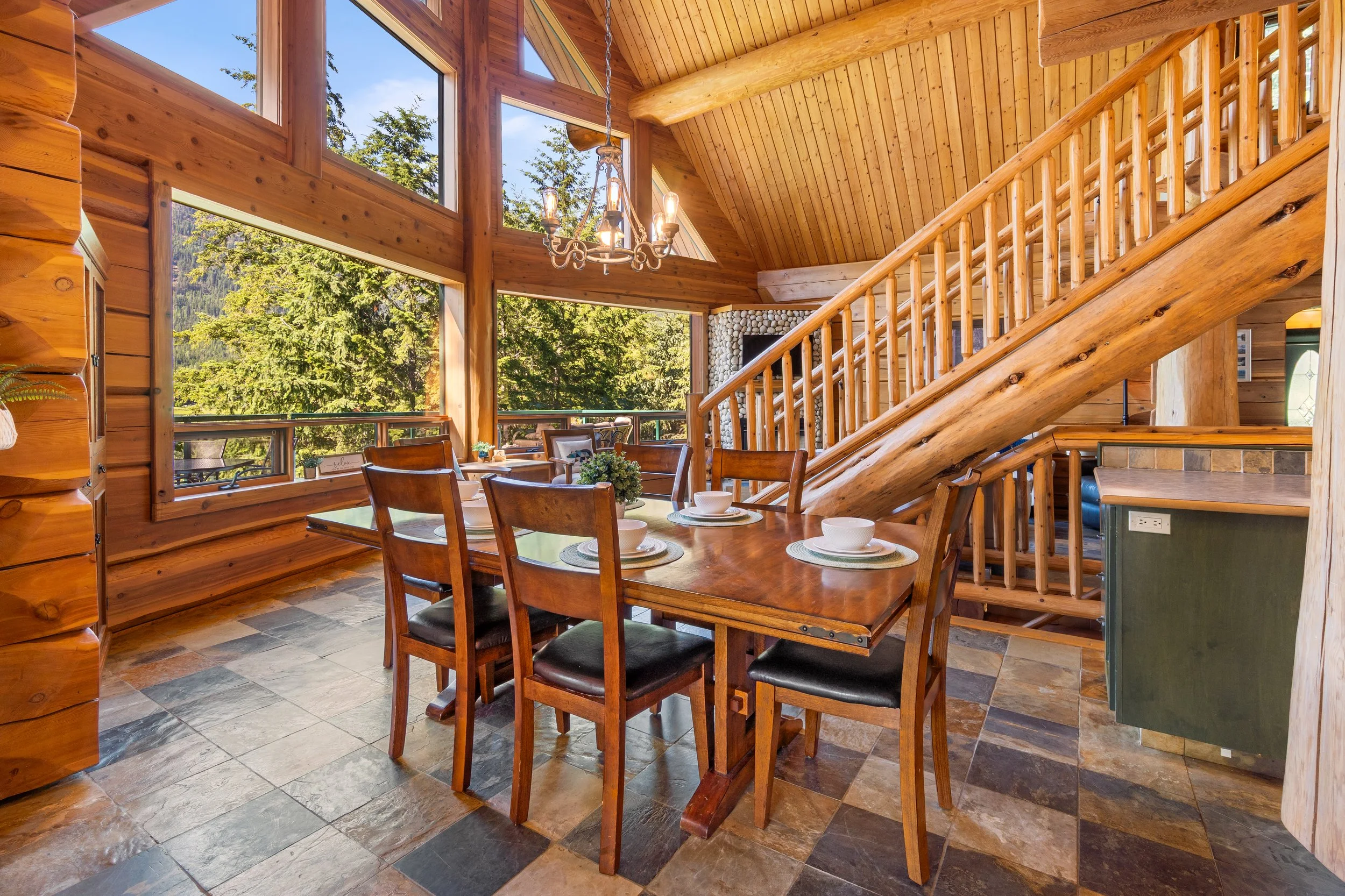 Interior of a log cabin dining area with large windows showing trees, wooden walls, a chandelier hanging above a wooden dining table set with bowls and placemats, and a staircase leading to an upper level.