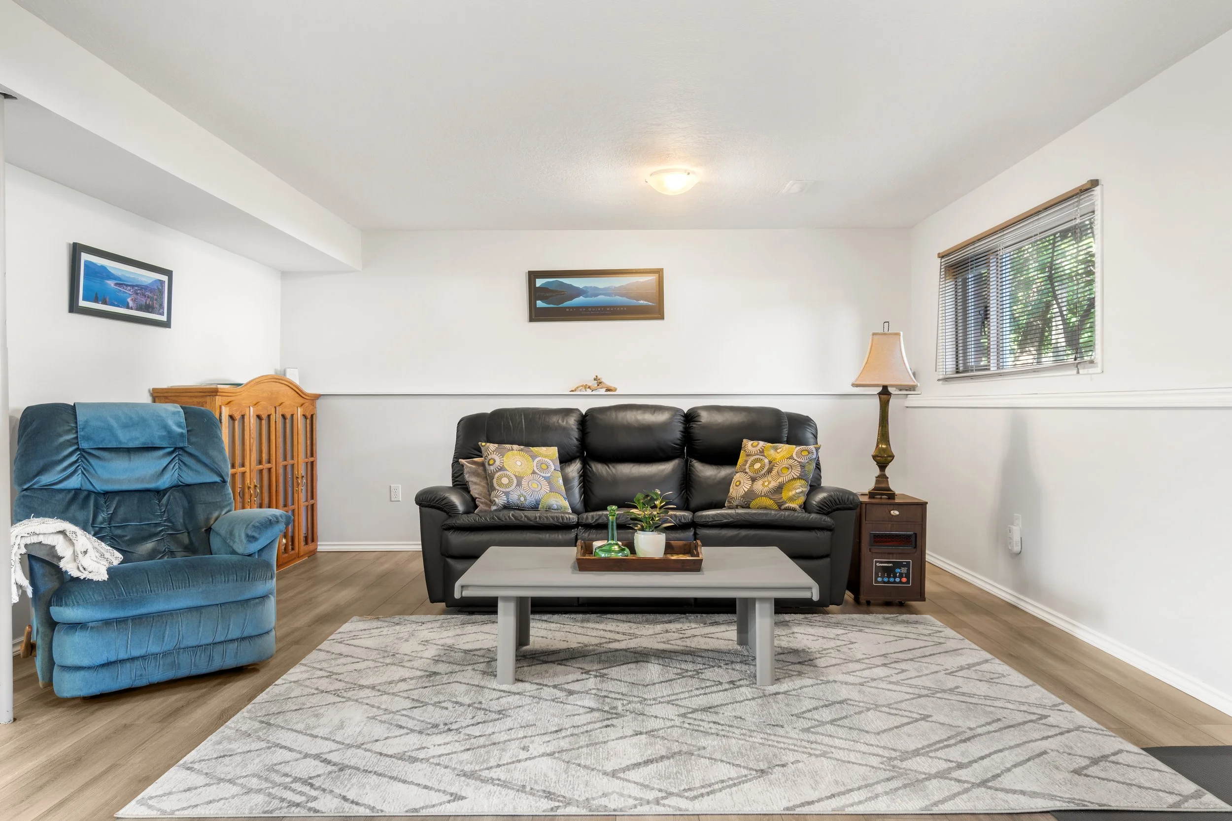 Living room with a brown leather sofa with patterned pillows, a blue armchair, a white coffee table with decorative items, framed landscape photos on the white walls, a window with blinds, a floor lamp, and a patterned rug on wood floors.