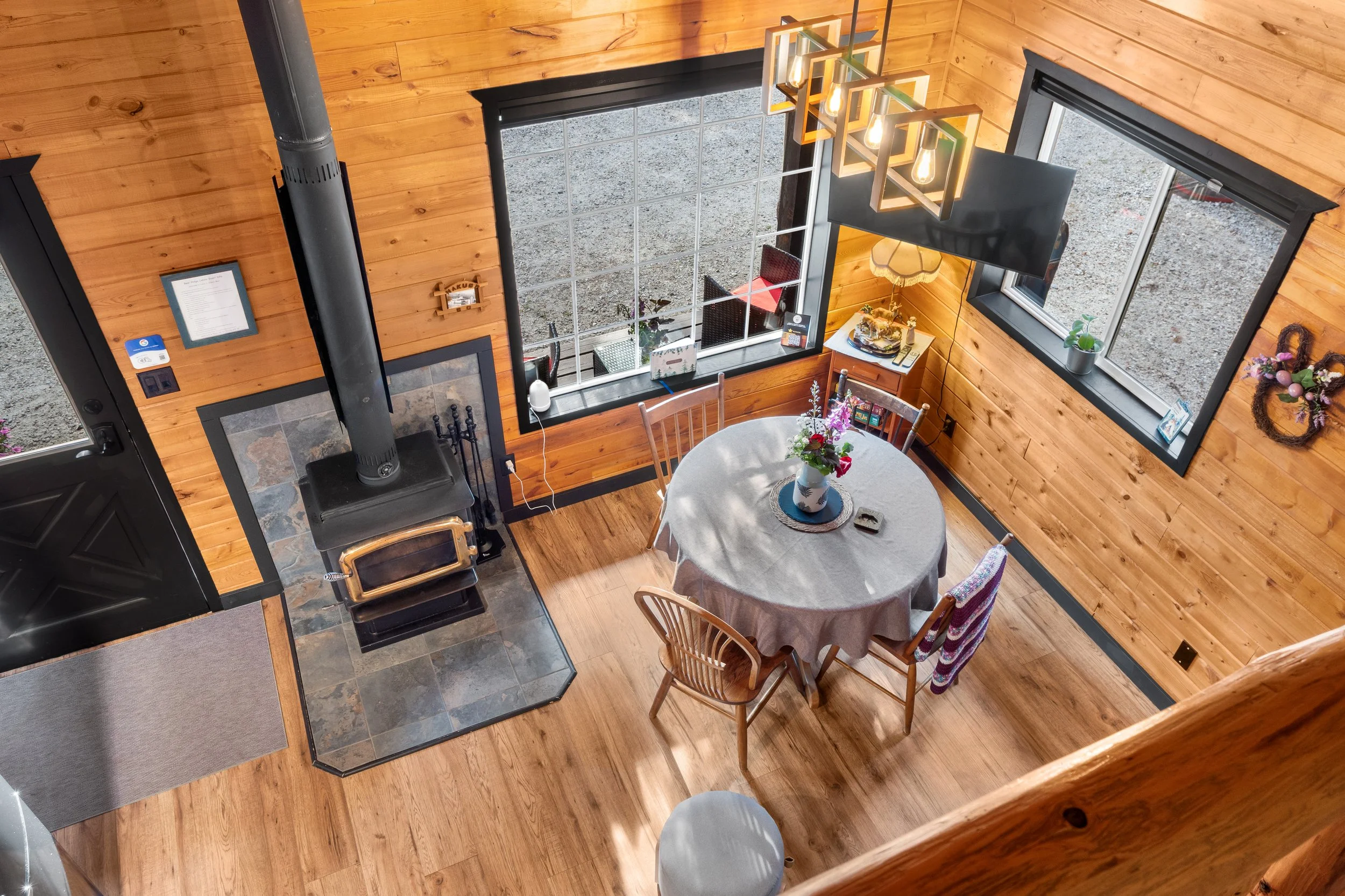 A cozy dining area with wooden walls, a round table covered with a tablecloth, and a vase with flowers. There are four wooden chairs around the table. A large window with black trim lets in natural light. A hanging light fixture with exposed bulbs is above the table. A wood stove is in the corner, next to a black firewood holder. The floor is wooden, and there's a gray doormat at the entrance.