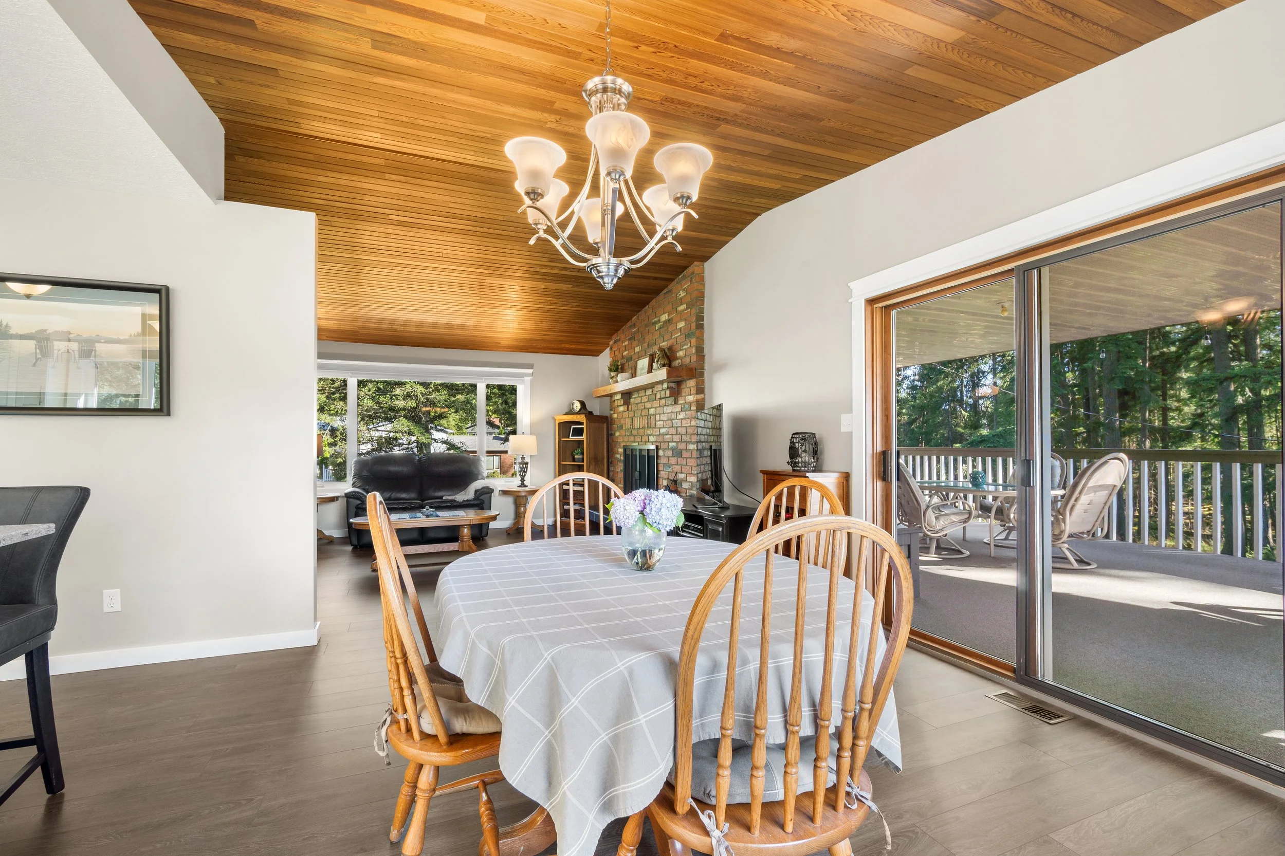 Interior view of a dining area with a round table covered with a plaid tablecloth, wooden chairs, a window with a view of trees, and a sliding glass door leading to a porch with outdoor furniture. The ceiling has wood paneling and a chandelier hanging above the table. There is a brick fireplace and a cabinet in the background.