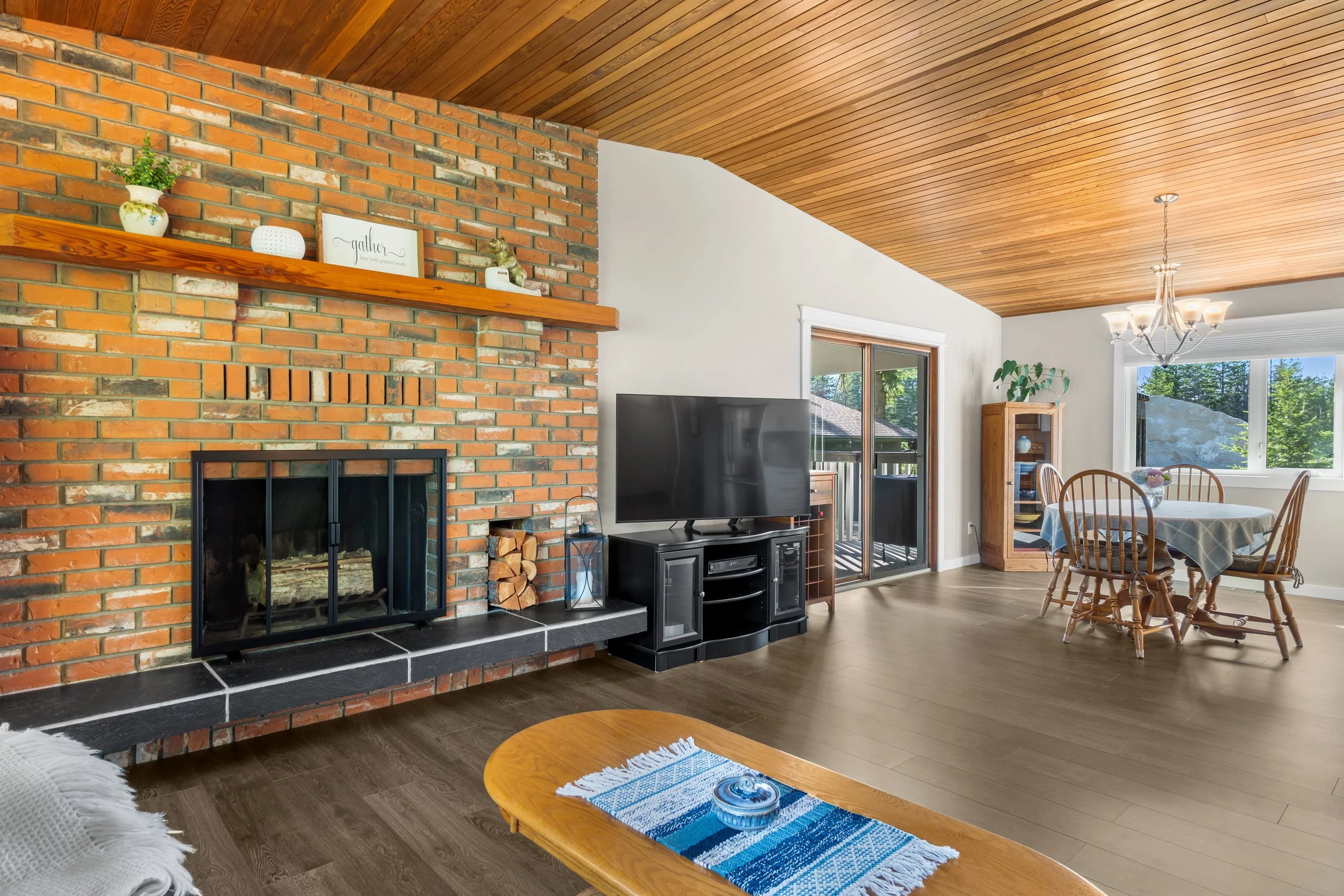 Living room with brick fireplace, wooden mantel, black TV stand, wooden dining table with chairs, window with view of trees, vaulted wooden ceiling, chandelier, and hardwood floors.