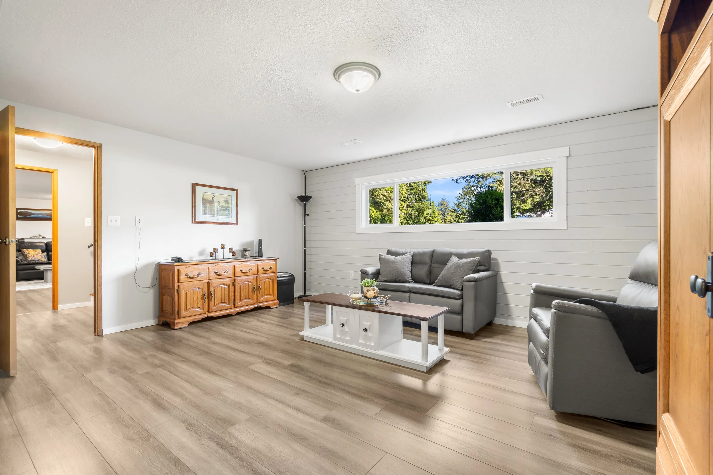 Living room with light wood flooring, white paneled wall, gray couches, wooden dresser, coffee table, and large window with trees outside.