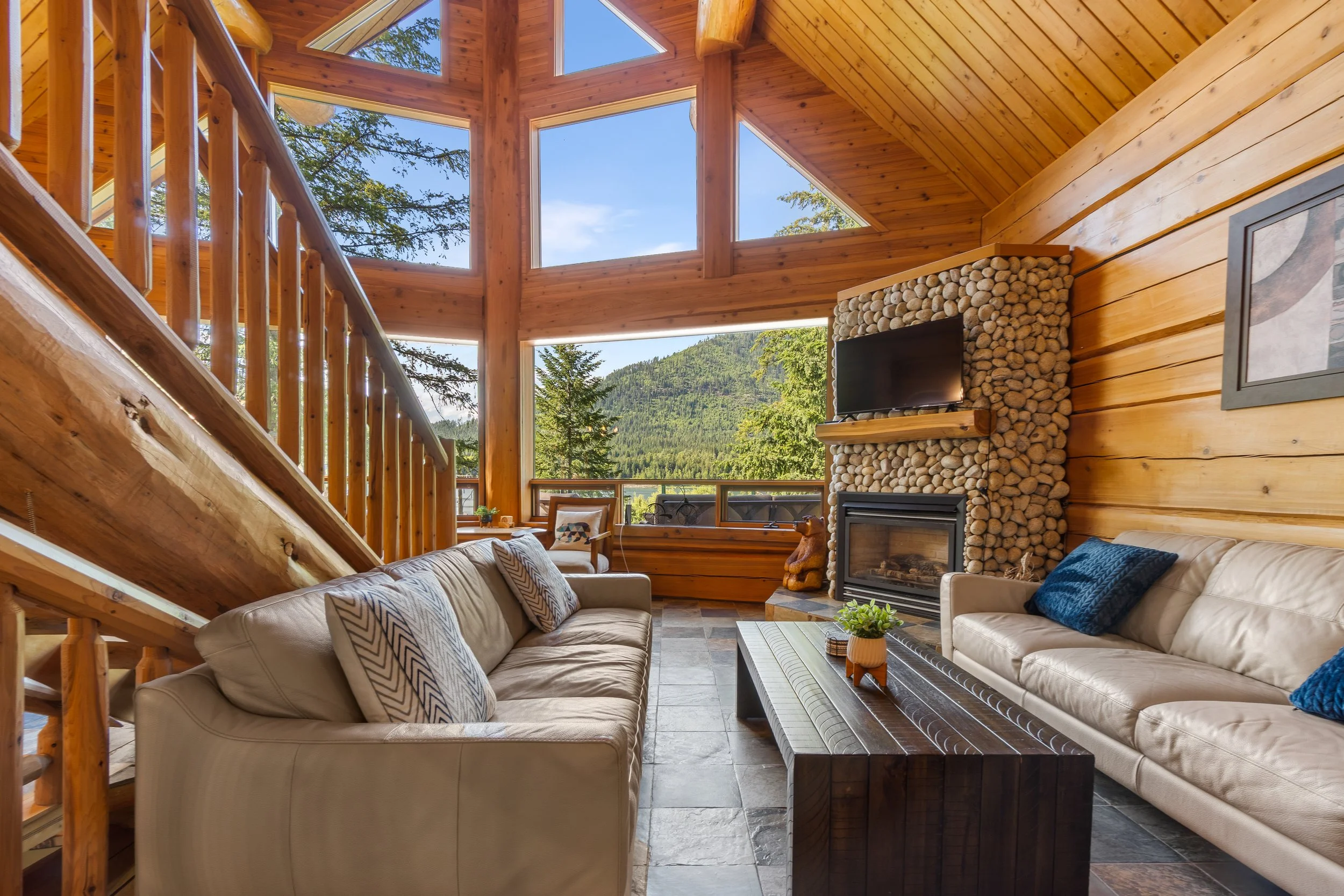 Living room with large windows showing mountains and trees, wooden walls and ceiling, stone fireplace with a flat-screen TV, beige sofas with patterned pillows, and a coffee table with a small potted plant.
