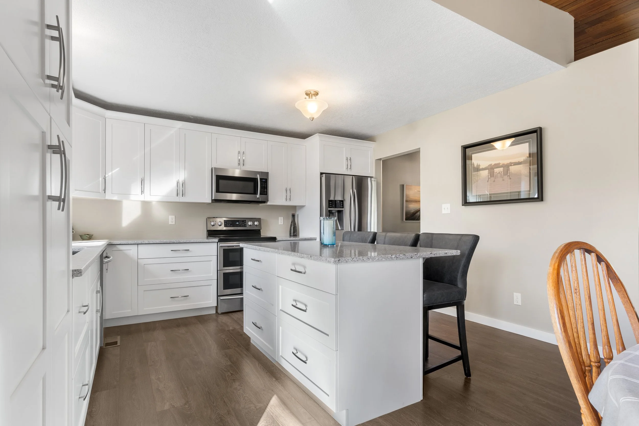 Modern kitchen with white cabinets, stainless steel appliances, and a large kitchen island with a granite countertop. There is a framed picture on the wall, a black chair, and part of a wooden dining table with a tablecloth.