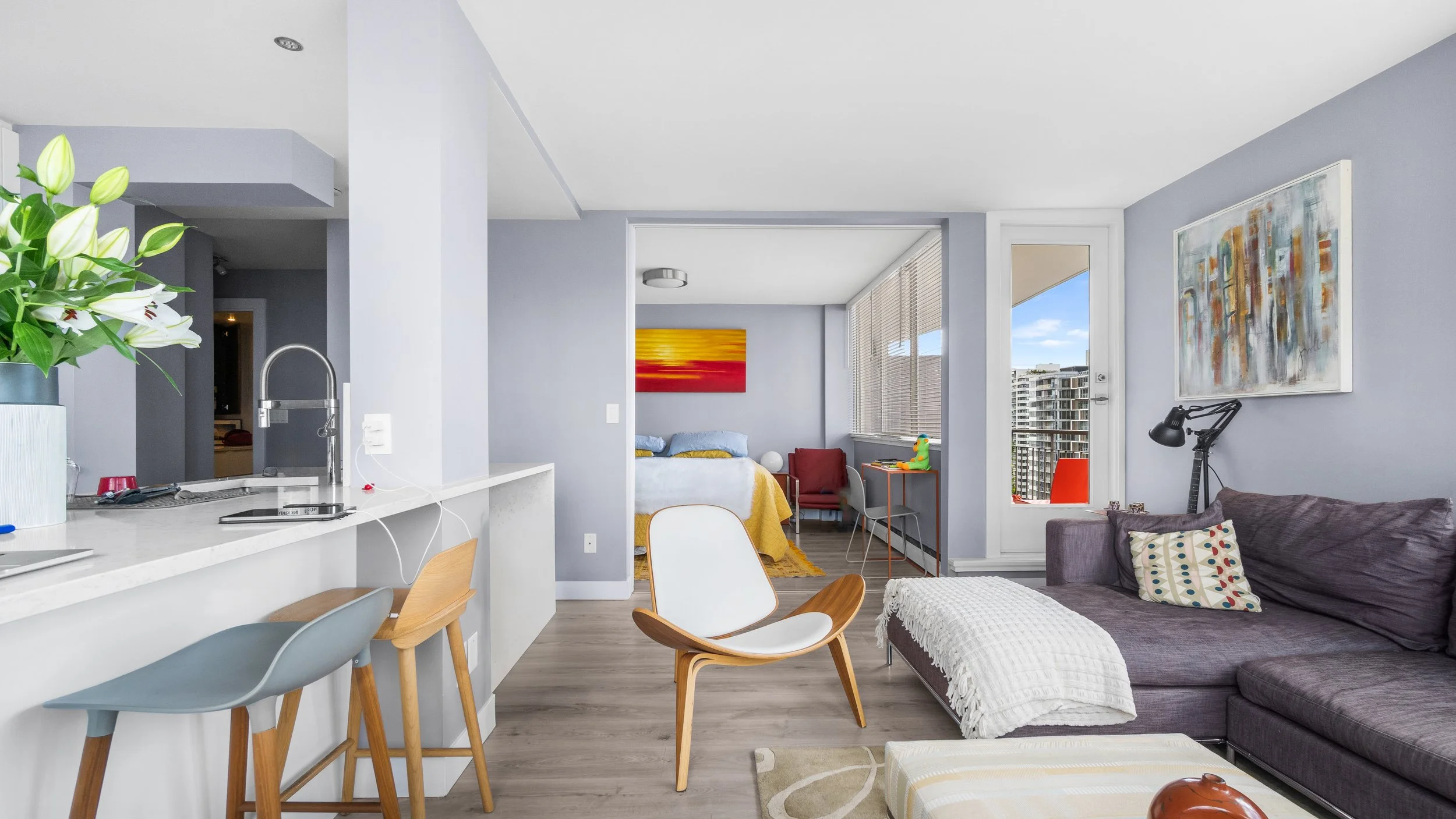 A modern apartment living room with a view into the bedroom, featuring a gray sofa with a patterned pillow, a white and wooden chair, and a white ottoman. The room has large windows with city views, colorful artwork on the walls, and a kitchen counter with a flower vase and barstools.