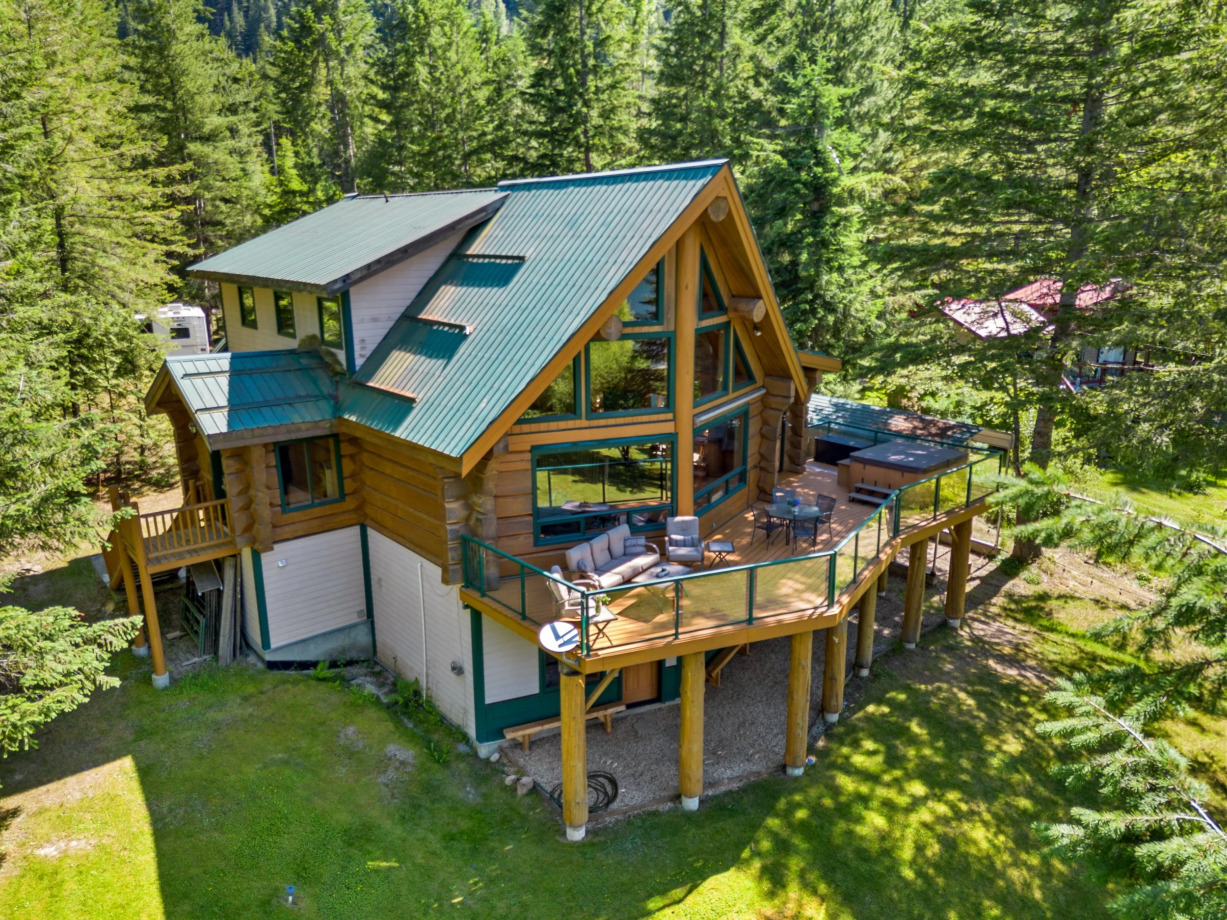 An aerial view of a wooden house with a green metal roof surrounded by tall pine trees in a forested area. The house has a large outdoor deck with seating and is elevated on stilts.