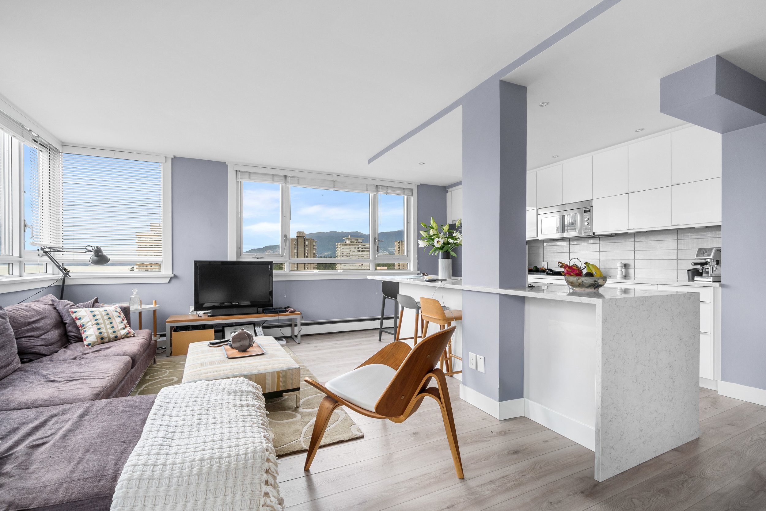 A modern open-concept living area with large windows showing city views, a purple sofa with pillows, a television, a coffee table, and a kitchen with white cabinets, a marble countertop, and a bowl of fruit.