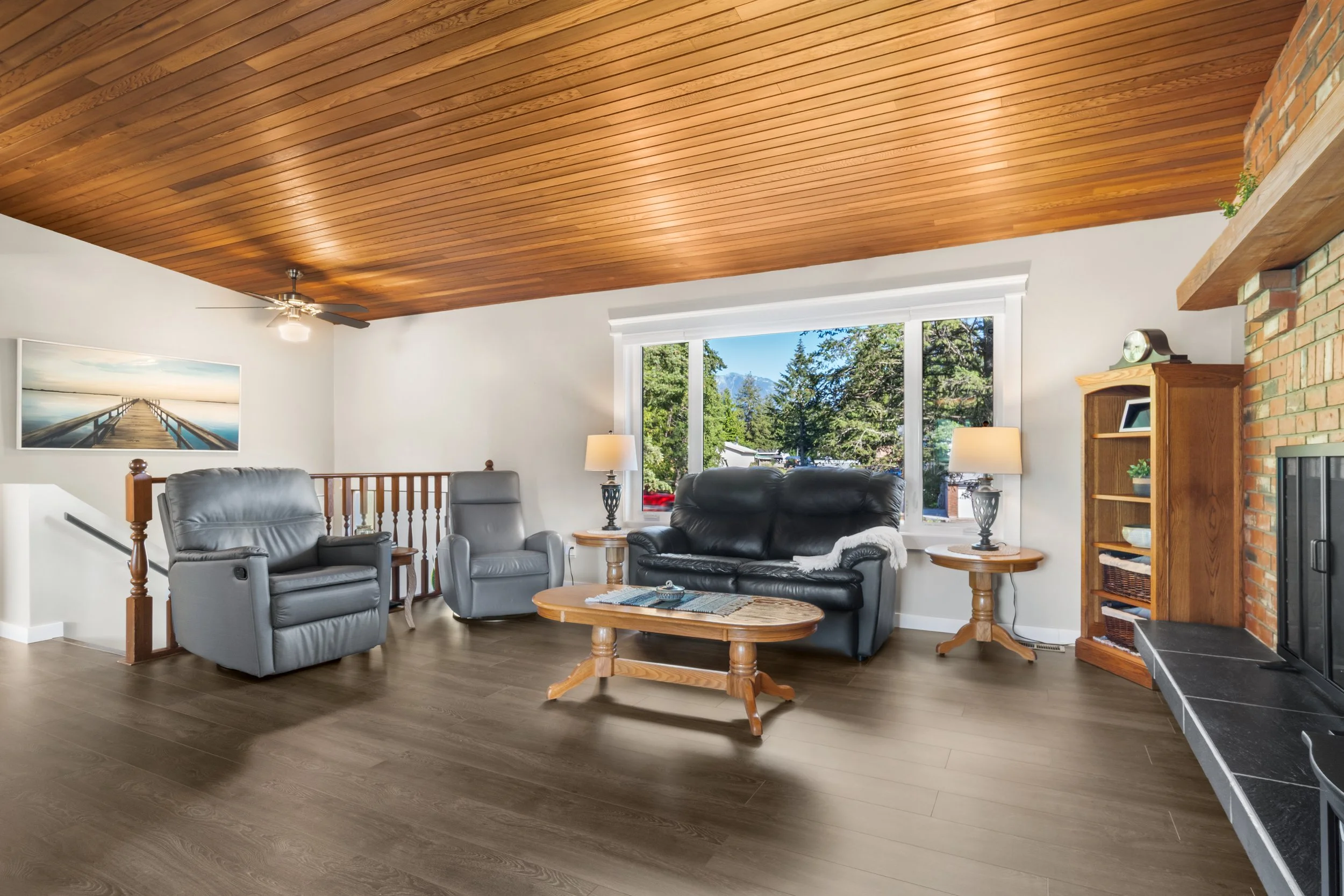 Living room with wood ceiling, large window showing trees and mountains, gray and black leather recliners, wooden tables, bookshelf, fireplace, and lamps.