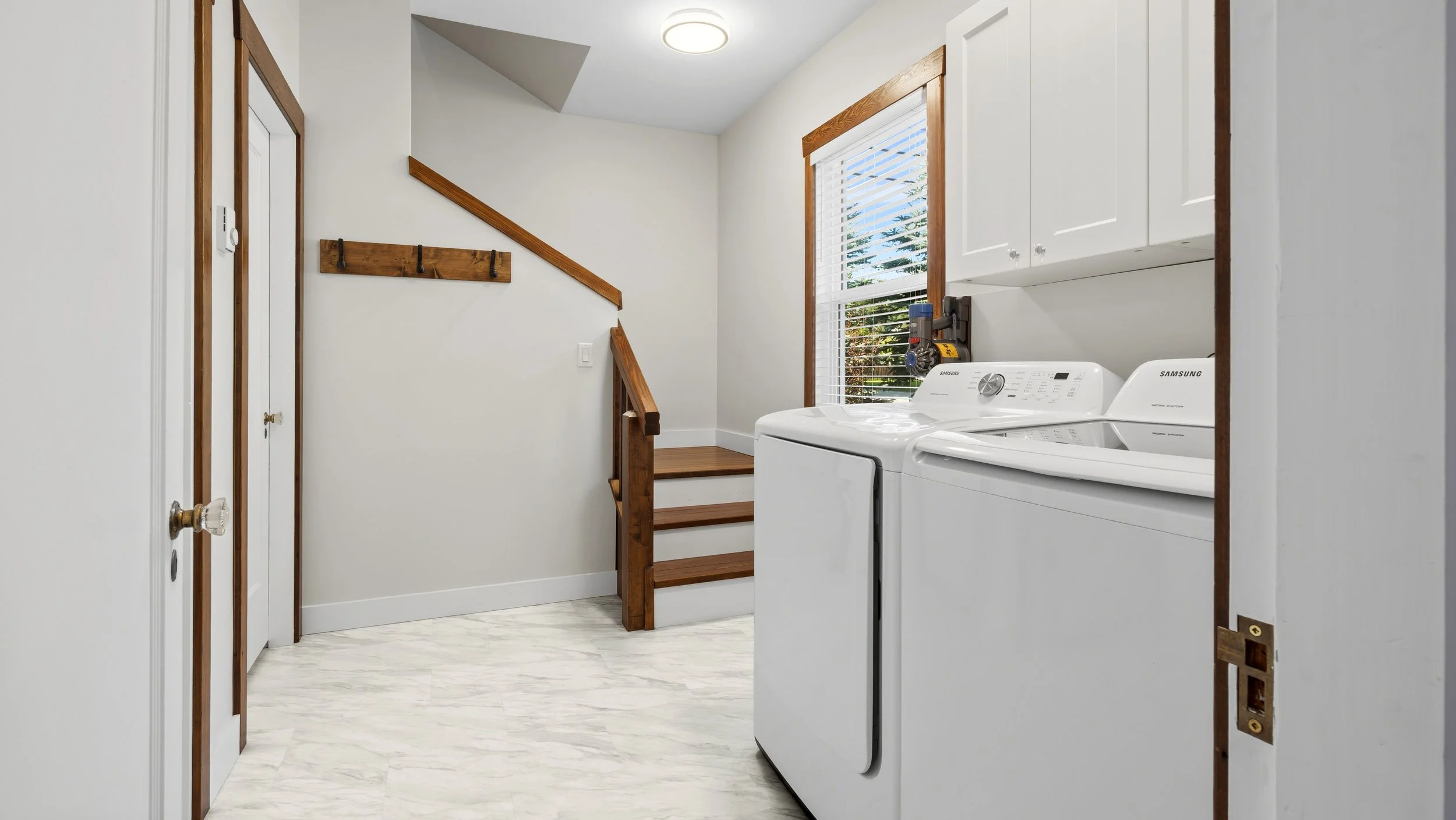A laundry room with white walls, a window with blinds, a white washing machine and dryer, and wooden accents on the trim and stairs. - Nakusp, 98 2nd Ave