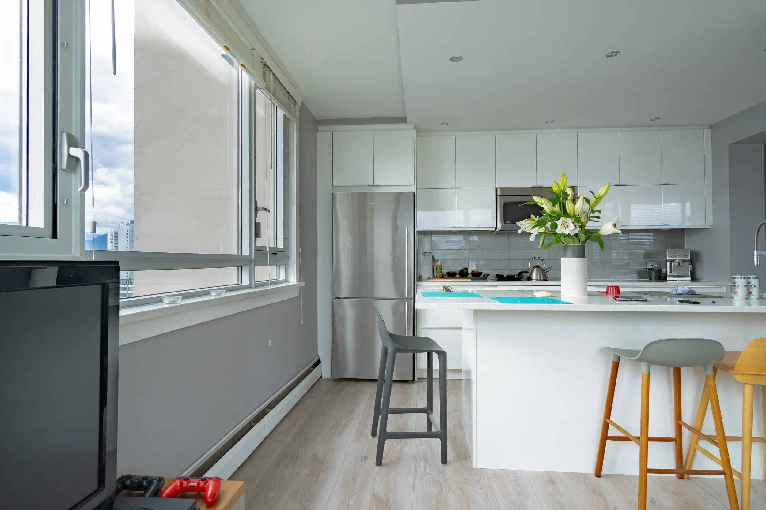 Modern kitchen with white cabinets, a stainless steel refrigerator, a microwave, a stove, and a vase with white lilies on the counter. There are large windows letting in natural light. Vancouver BC, 1903 Comox St.