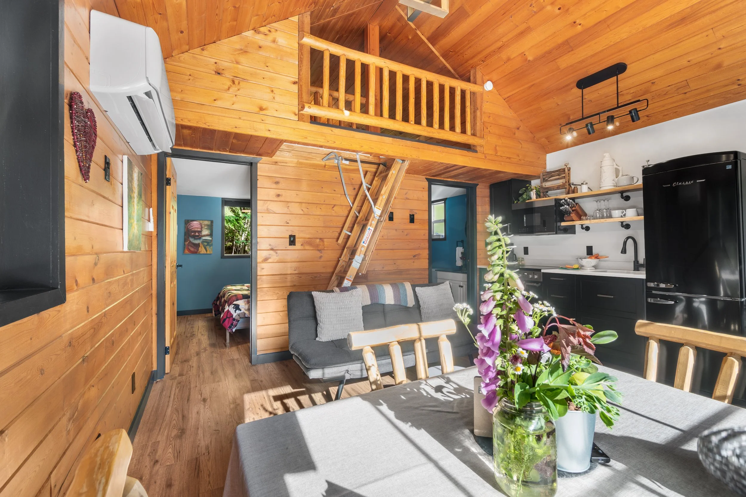 Interior of a kitchen and living room with wooden walls and ceiling, featuring black appliances, open shelving with dishes, a gray sofa, a dining table with a flowering plant centerpiece, and a loft area with a wooden railing.
