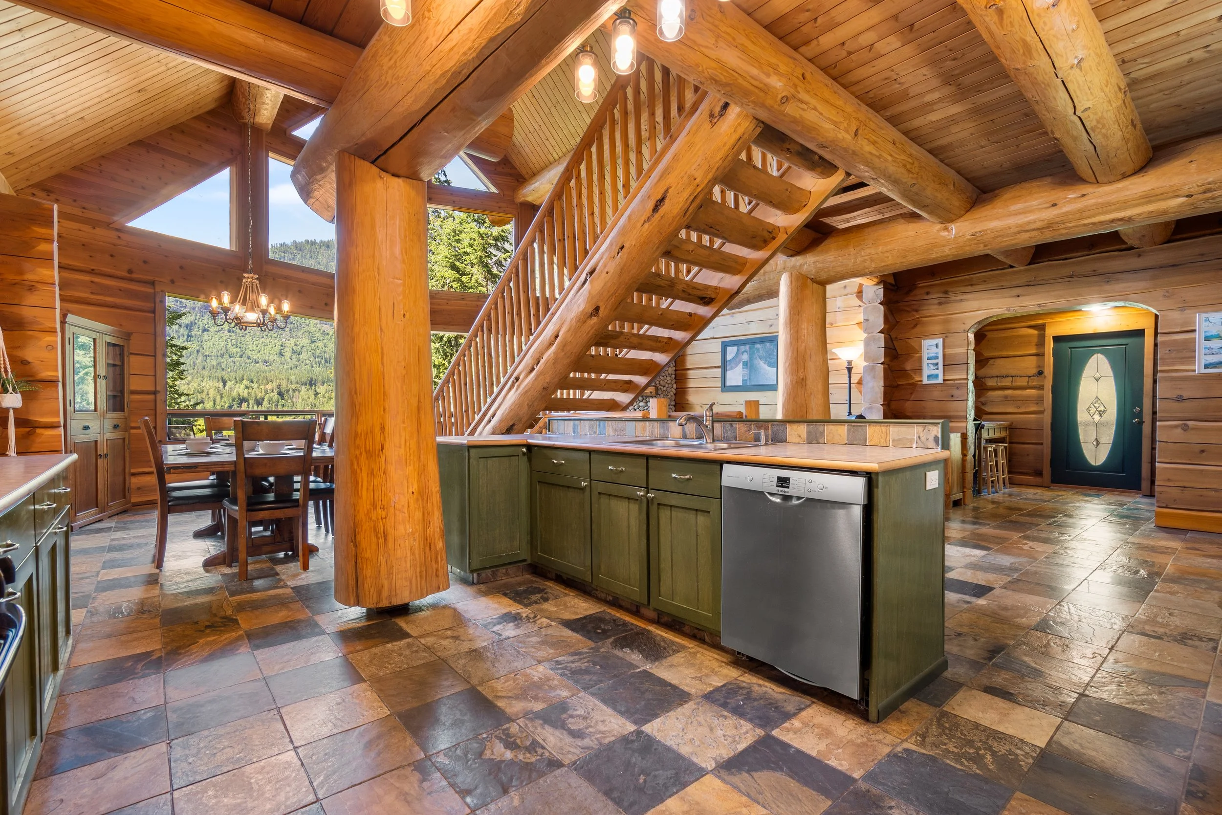 Interior of a log cabin with a wooden staircase, a green kitchen island, a dining table, and large windows showing trees and mountains outside.