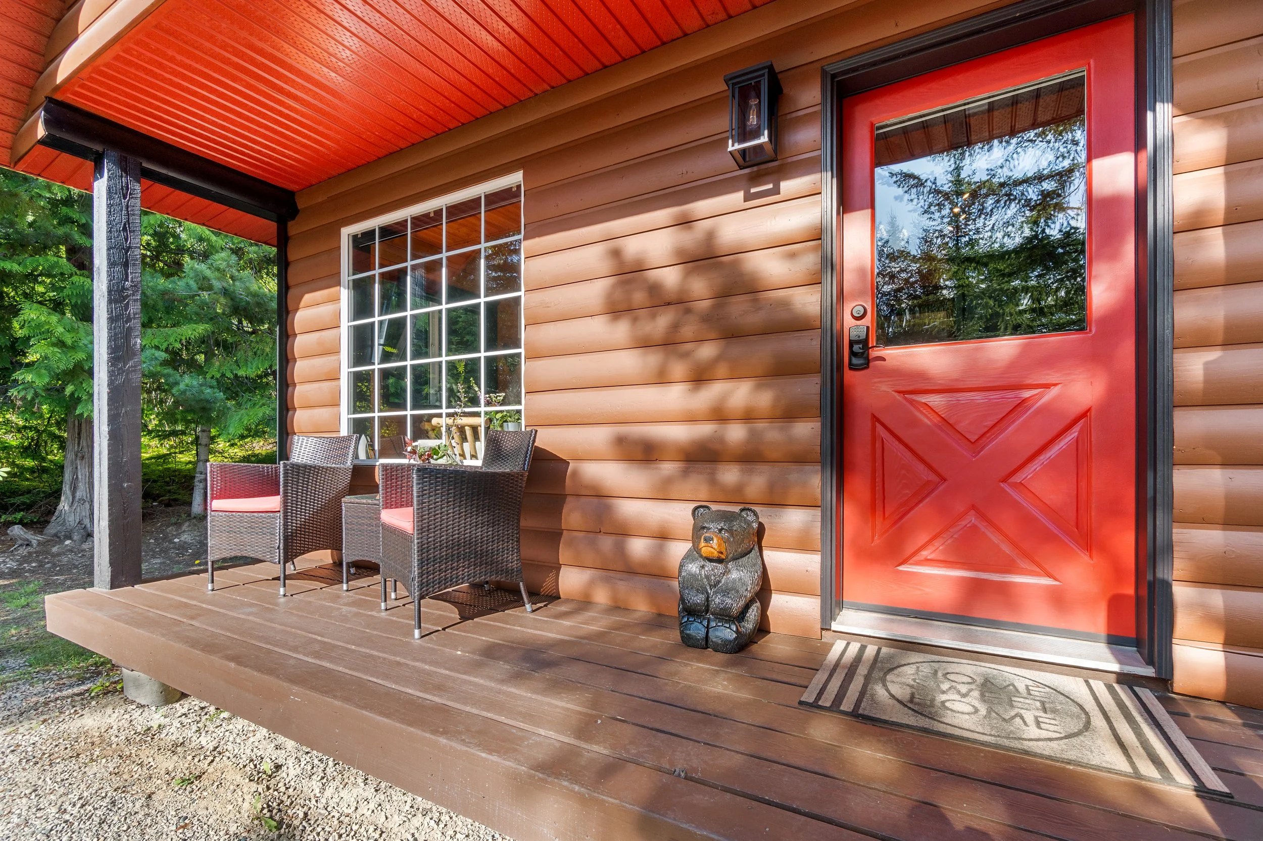 A porch of a wooden house with a red door, a large window, and outdoor seating with chairs. There is a teddy bear statue next to the door and a welcome mat on the deck.