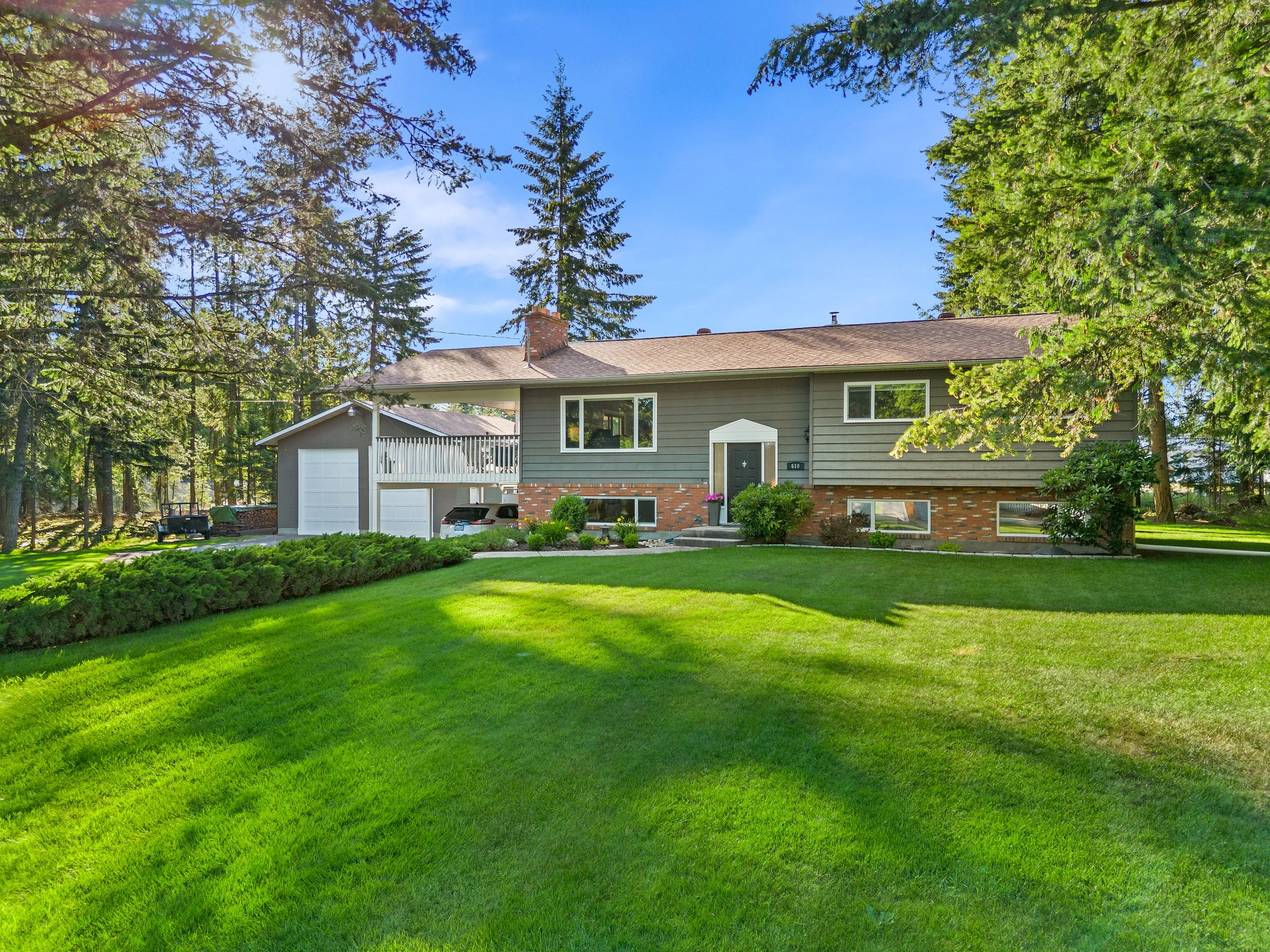 A two-story house with gray siding and brick base, surrounded by green lawn and trees, under a clear blue sky.