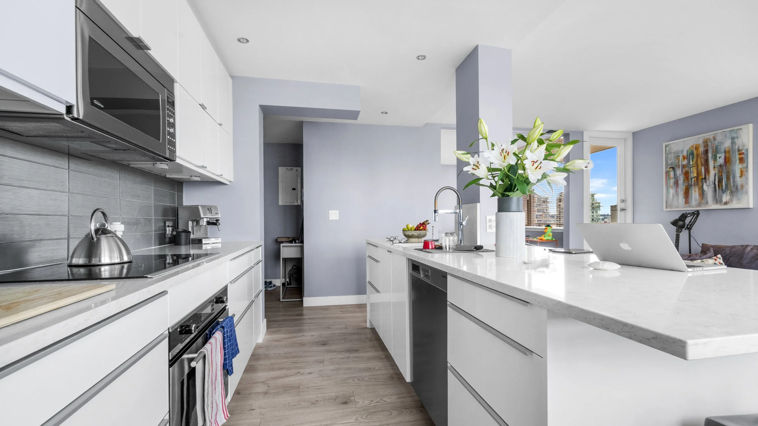 Modern kitchen with white cabinets, marble countertops, a stove with a tea kettle, and a large window with city views, decorated with flowers and artwork.