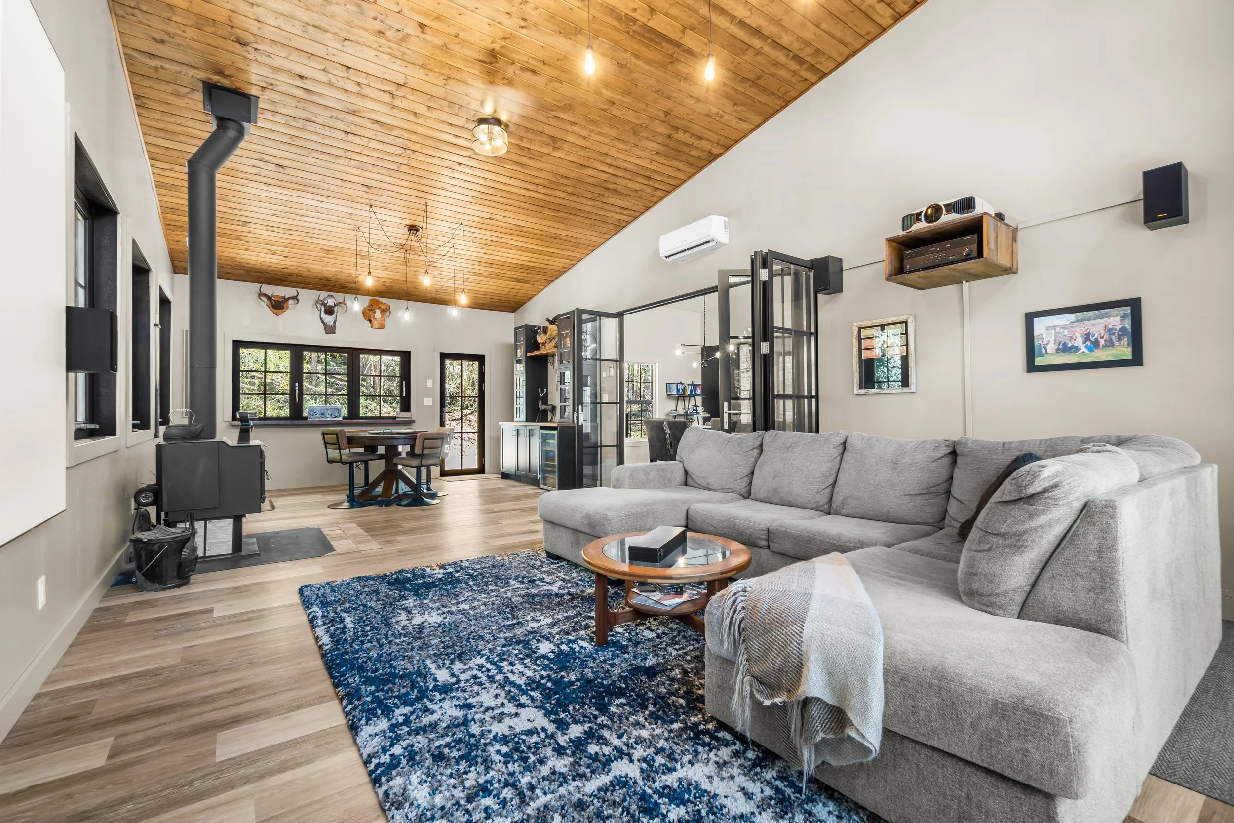 Modern living room with a gray sectional sofa, a round wooden coffee table, a blue patterned rug, wood ceiling, multiple windows, and a dining area in the background with a round table and chairs. - Trail BC Real Estate Photography