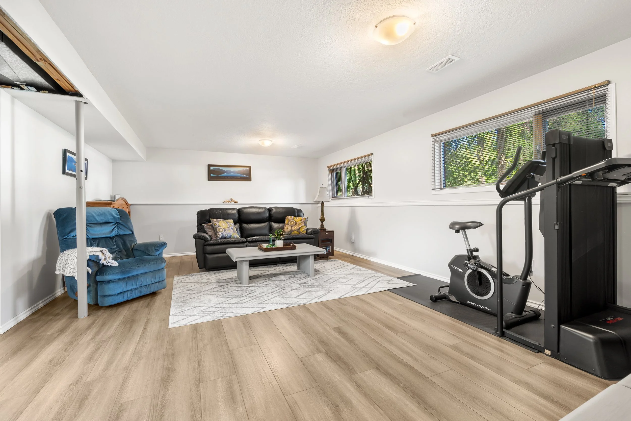Living room with a black leather sofa, blue armchair, white coffee table, exercise bike, large window with blinds, small framed landscape picture on wall, and wood flooring.