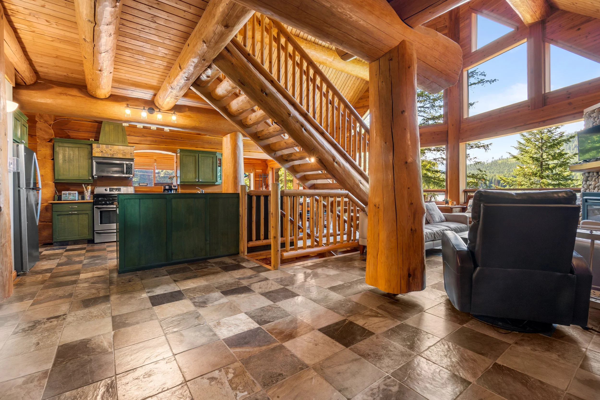 Interior of a log cabin with a open living space, visible staircase made of logs, large windows showing trees and a blue sky, kitchen area with green cabinets, patterned tile floor, and a seating area with sofa and chairs.
