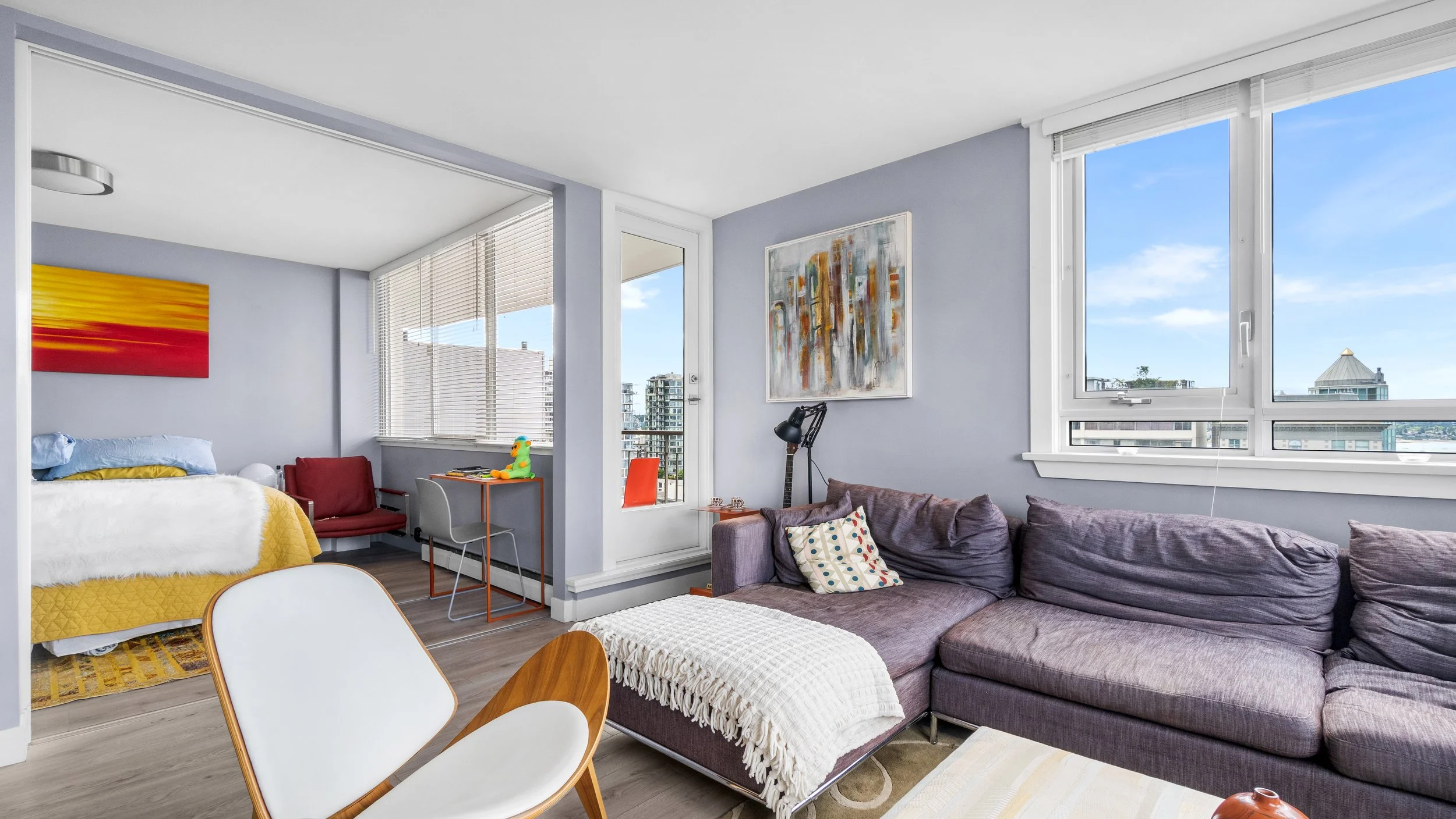 Interior view of a modern apartment living room with large windows, a purple sofa with pillows, a white armchair, abstract wall art, and a bedroom area with a yellow blanket and colorful artwork.