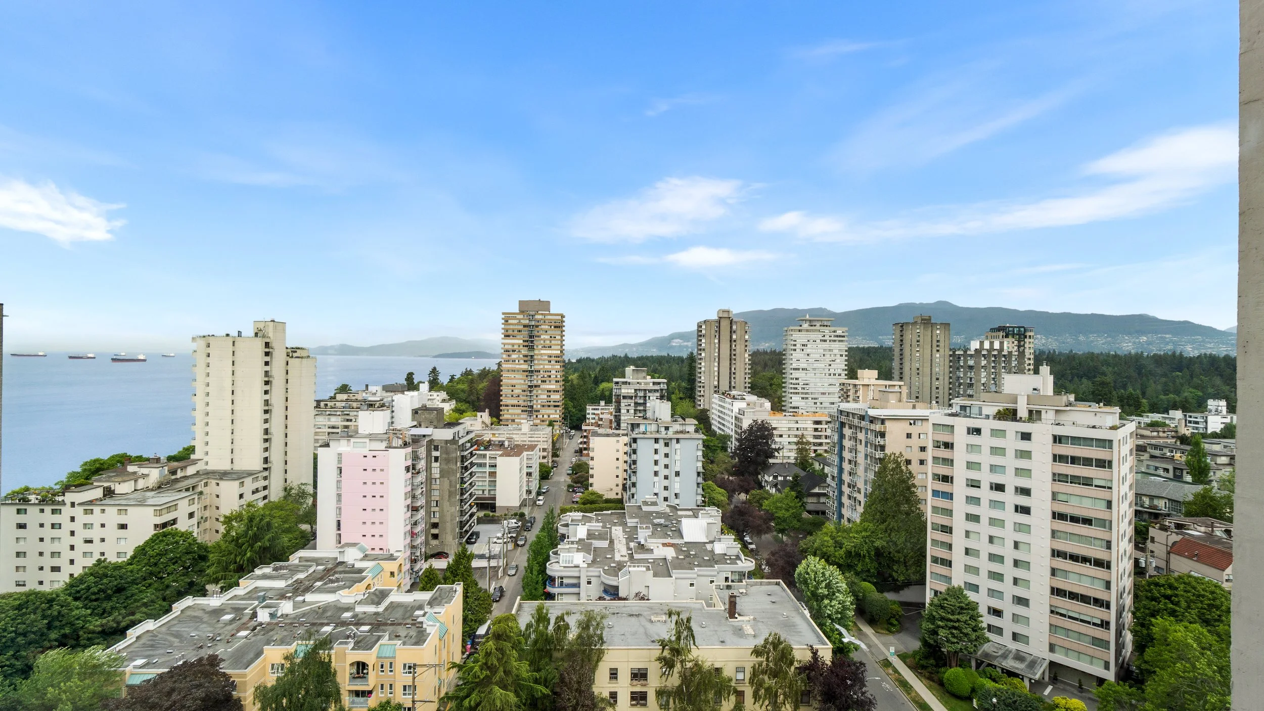 View of a cityscape with high-rise buildings and trees, overlooking a body of water with ships, under a partly cloudy sky.