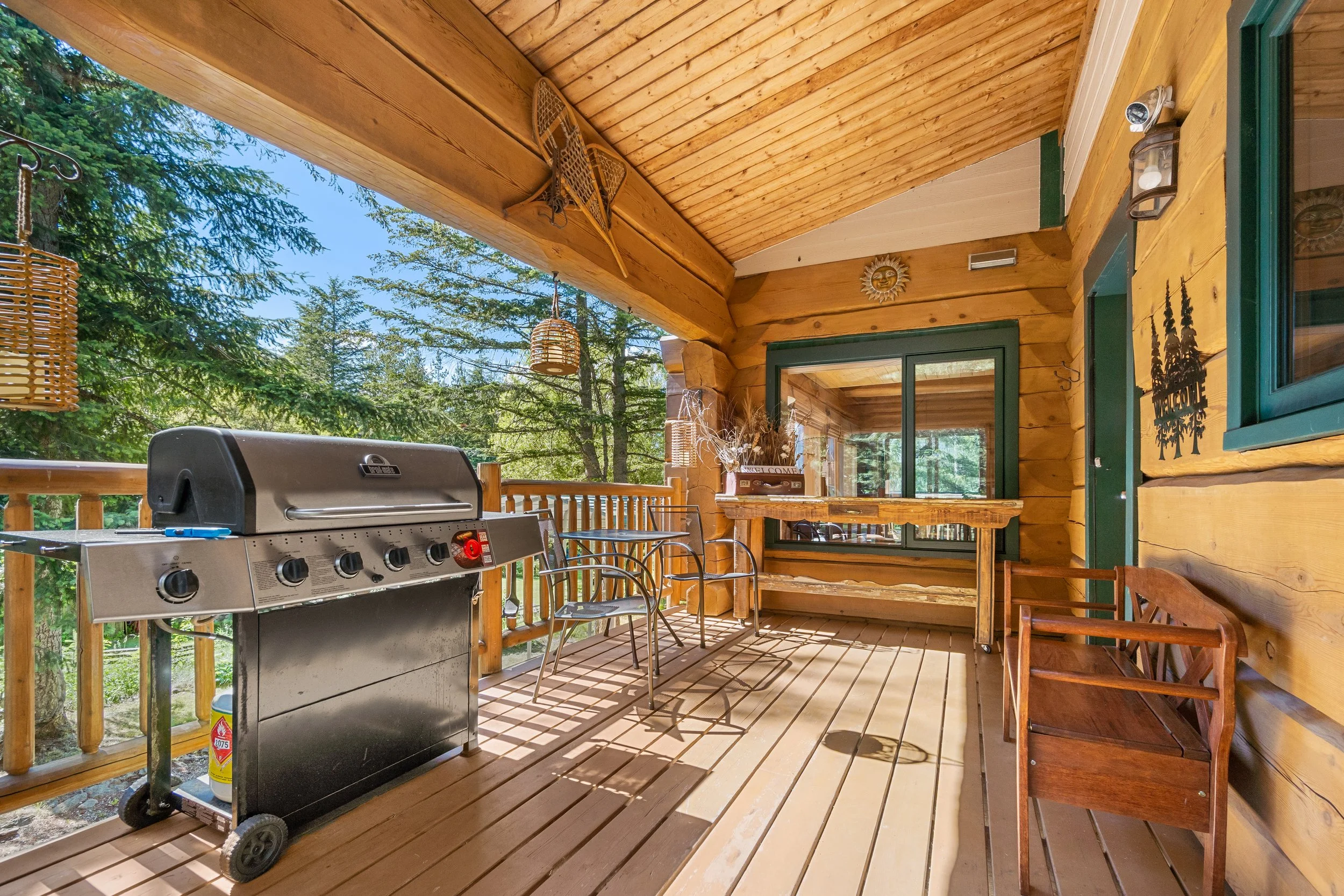 A covered wooden porch with a stainless steel gas grill, metal chairs, and wooden benches, surrounded by trees and greenery.