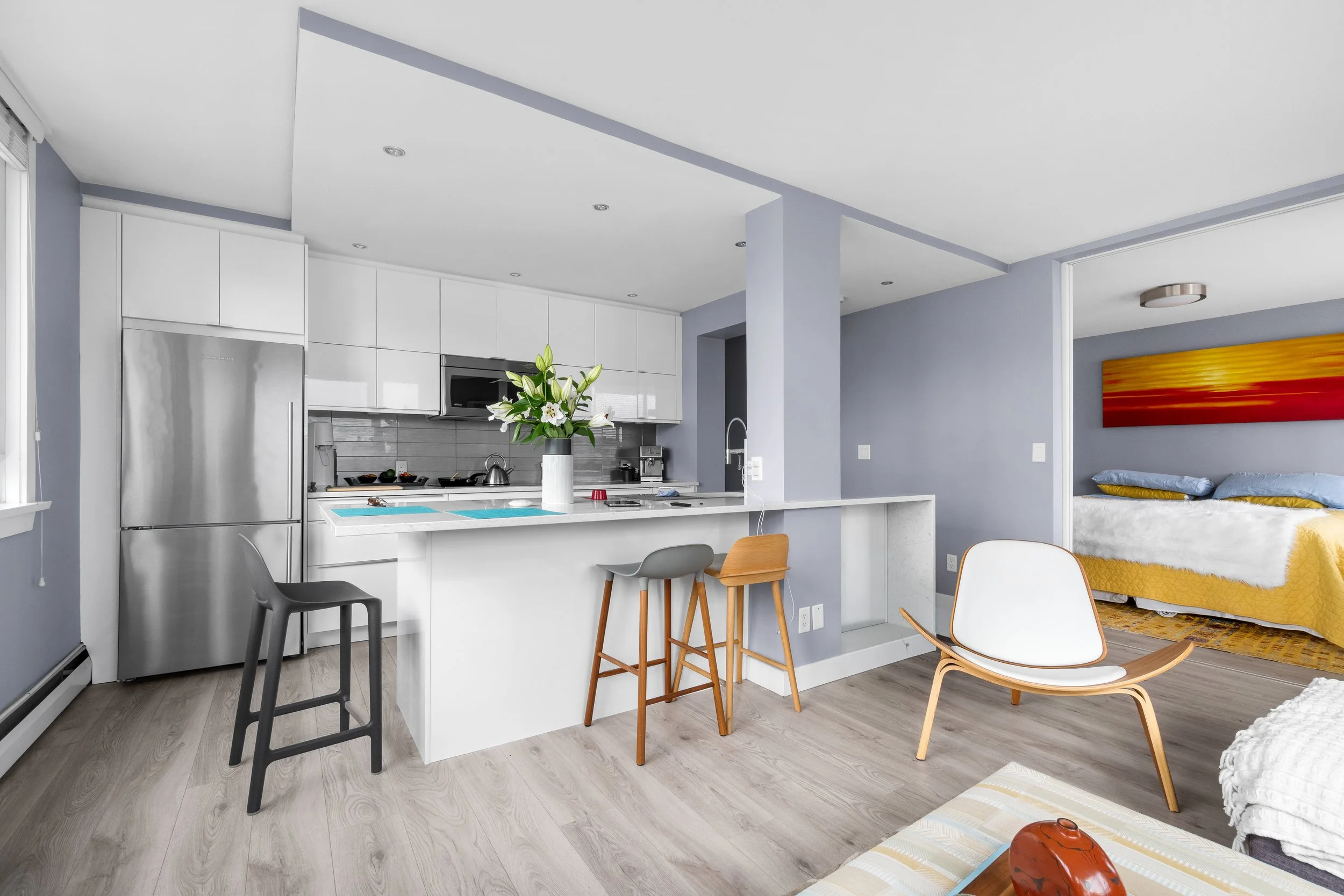 A modern open-plan kitchen and living area with light wood flooring, white cabinetry, a flower vase on the kitchen island, and colorful wall art in the background.