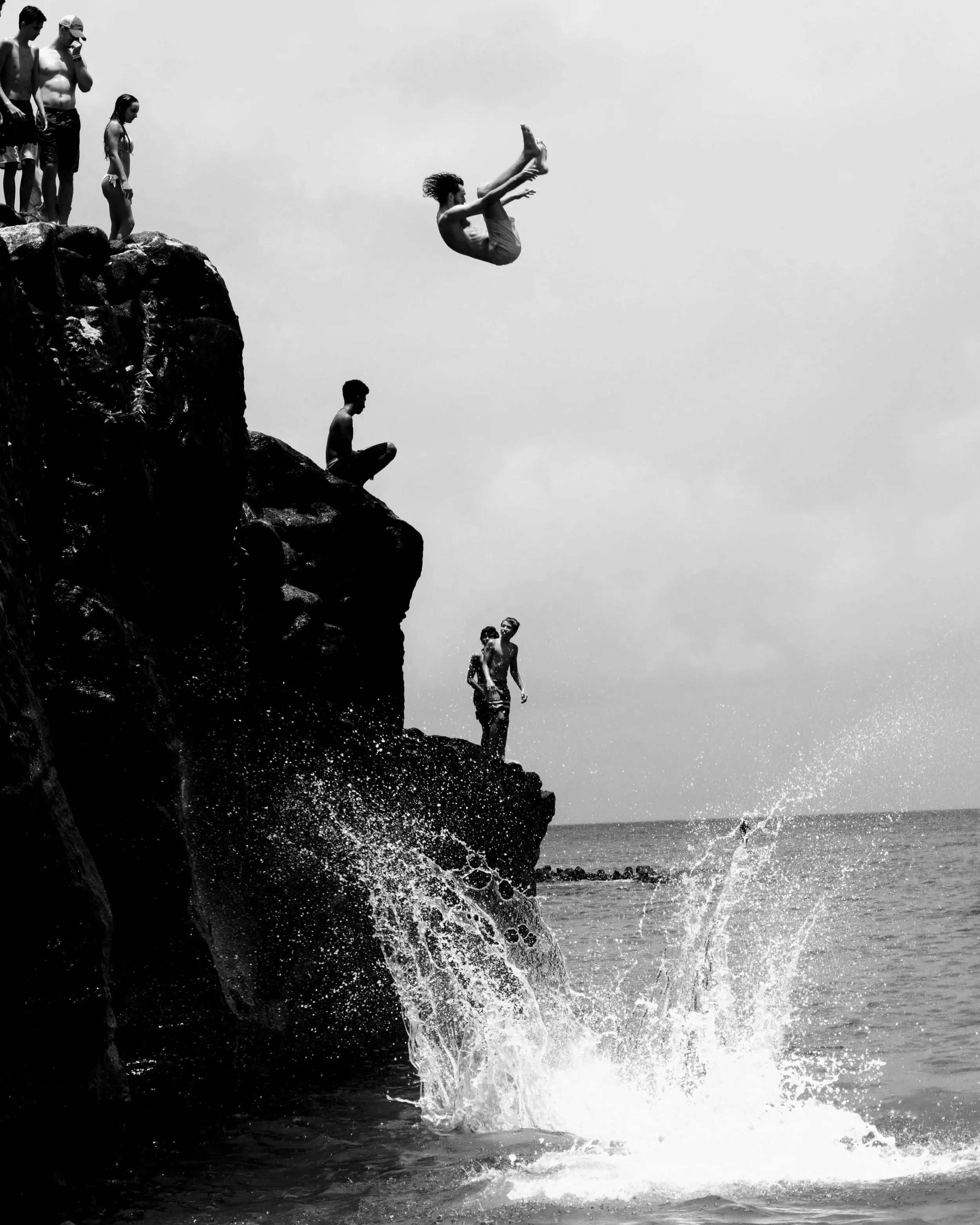 Black and white photograph of people cliff diving into water, with one person suspended midair above a large splash, evoking risk, aliveness, emotional intensity, courage, pleasure, and the full experience of being human.