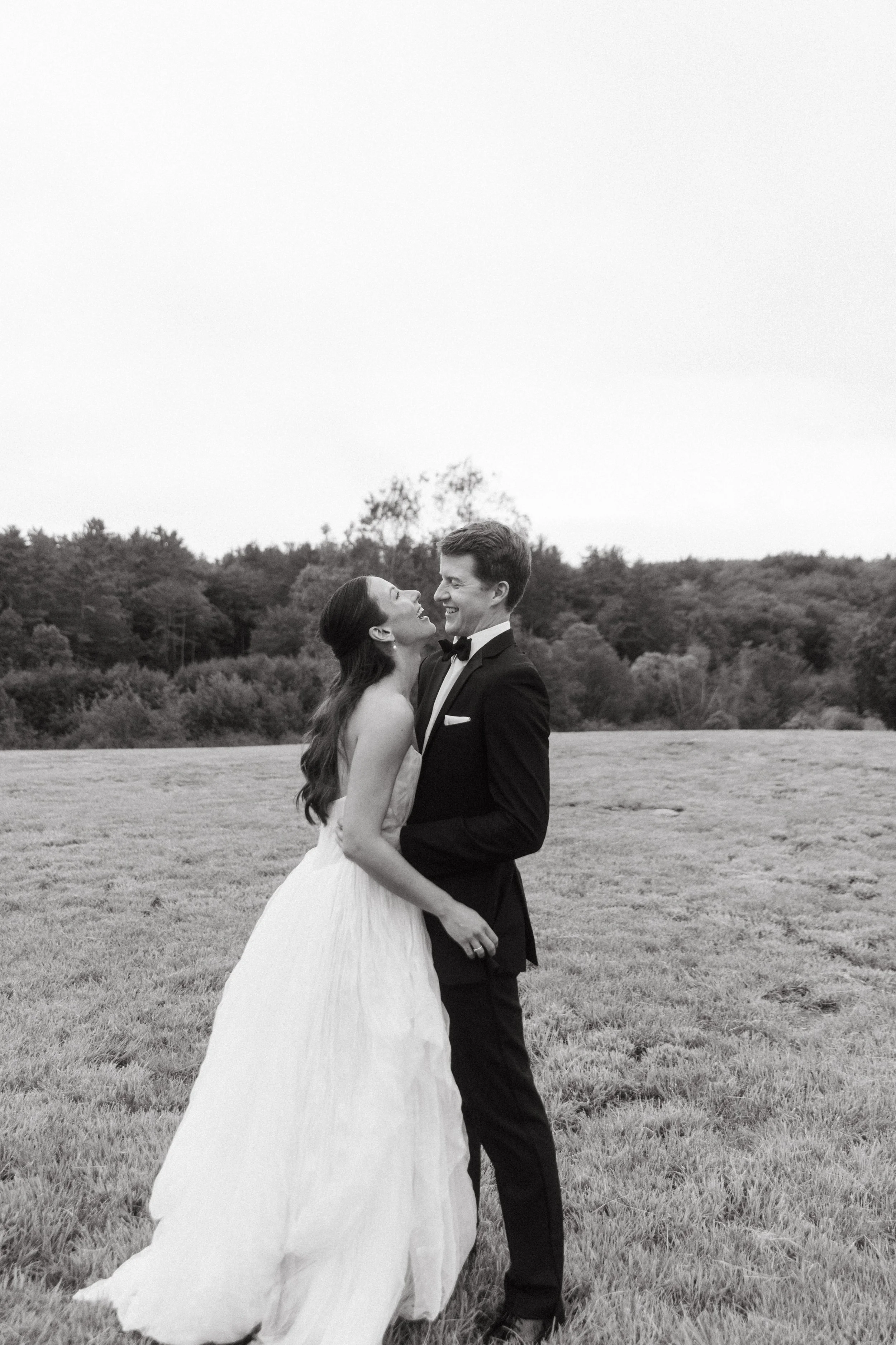 A black and white photo of a bride and groom standing in a grassy field, smiling and looking at each other, with trees in the background.