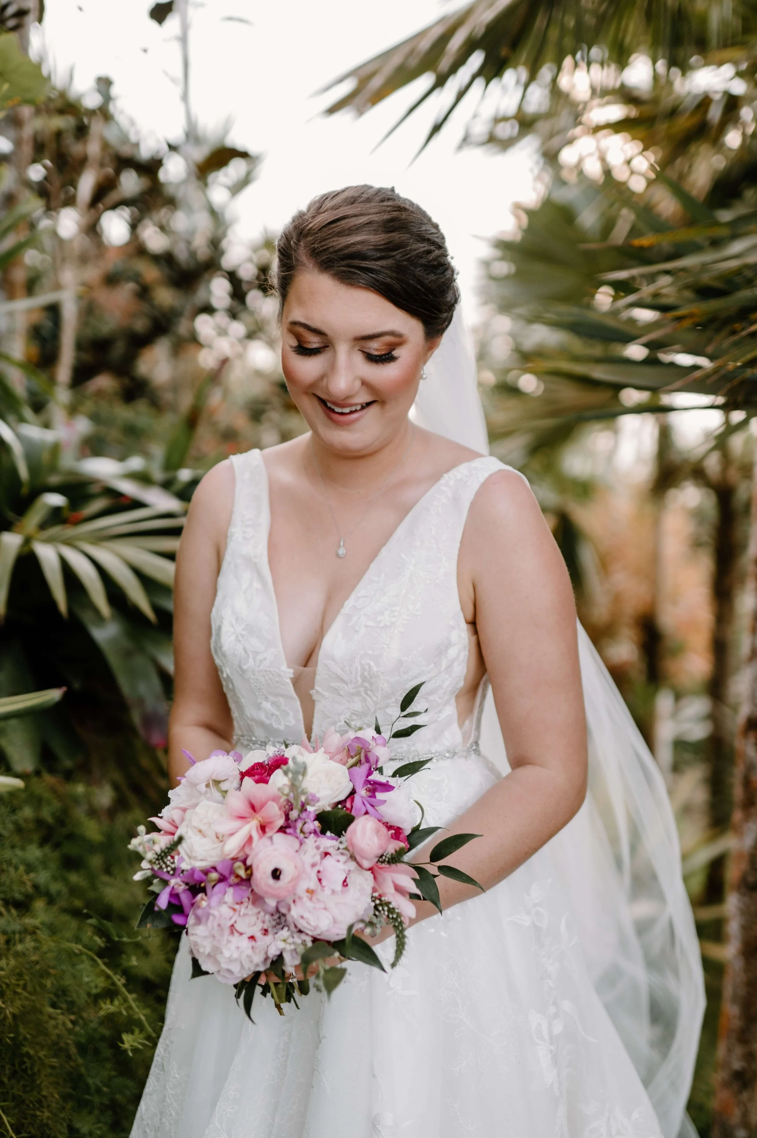 Bride in a white wedding dress holding a pink and white floral bouquet outdoors surrounded by lush green plants and trees.