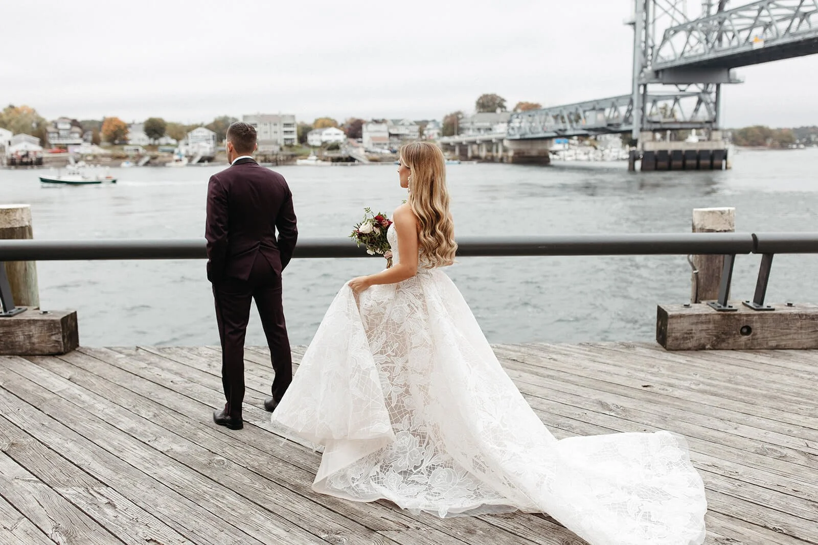 Bride in a white lace wedding gown holding a bouquet, standing on a wooden dock by water, with groom in a dark suit facing away, and a bridge in the background.