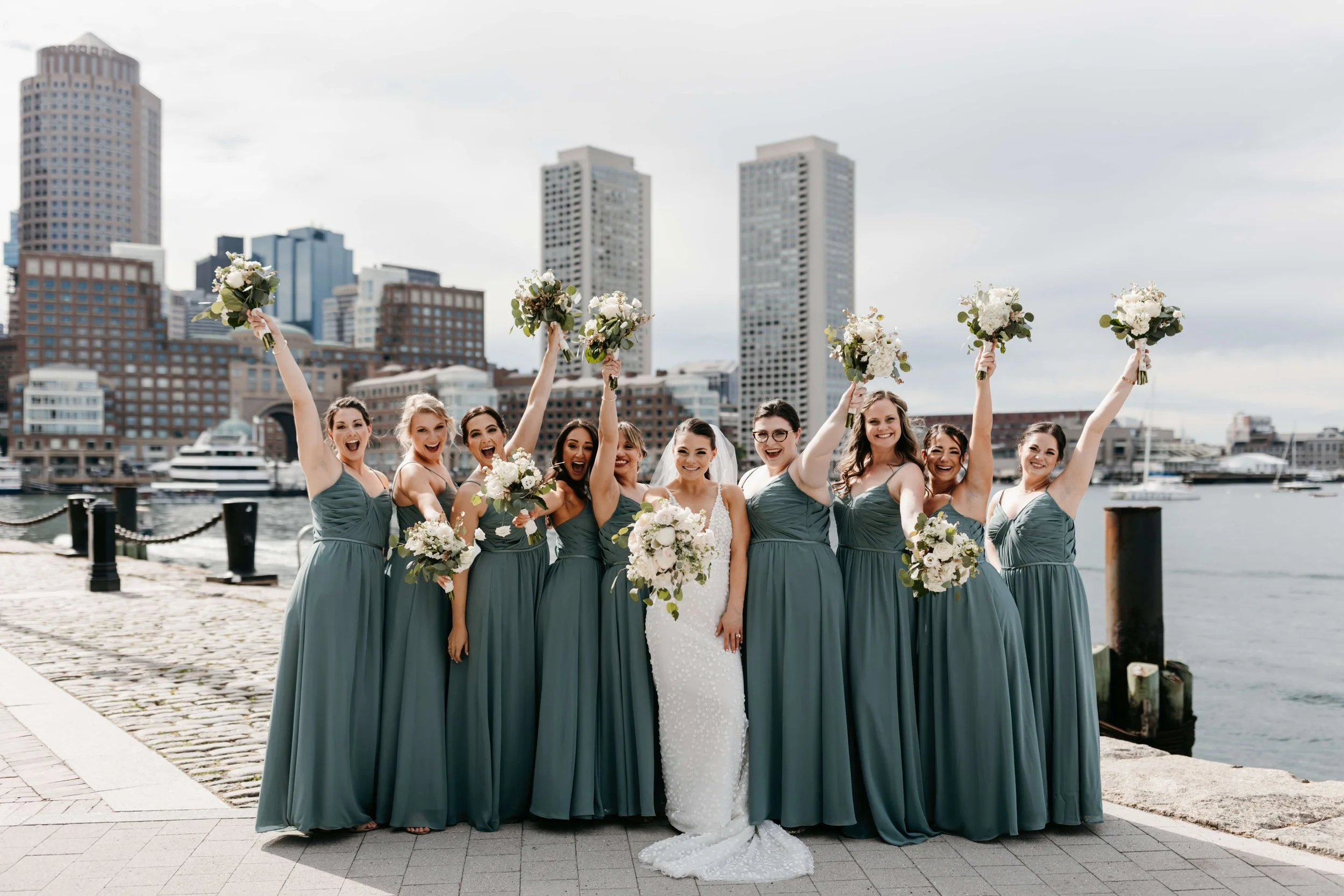 A bride and nine bridesmaids celebrating by the waterfront with city buildings in the background, holding bouquets and smiling.