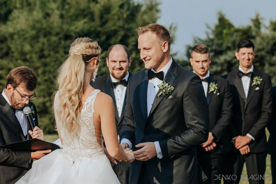 A bride and groom holding hands during an outdoor wedding ceremony, with groomsmen standing behind them, under a bright sky with trees in the background.