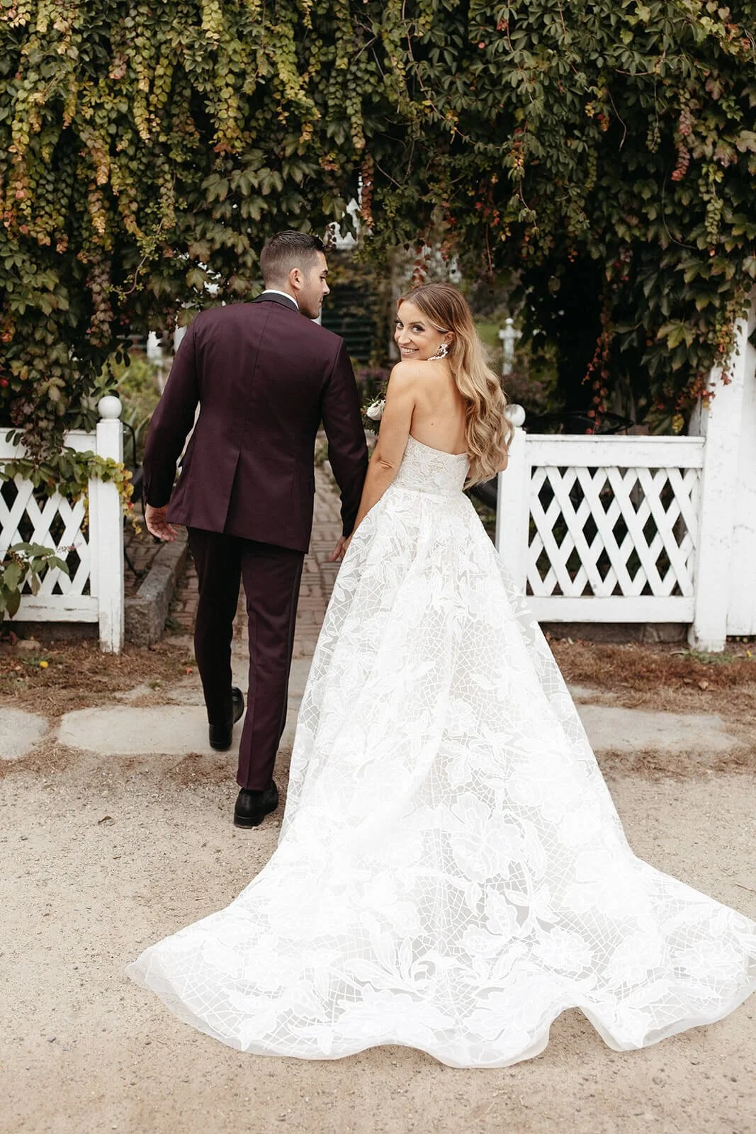 Bride and groom holding hands and walking through a garden gate, the bride smiling back at the camera, with lush green foliage overhead.