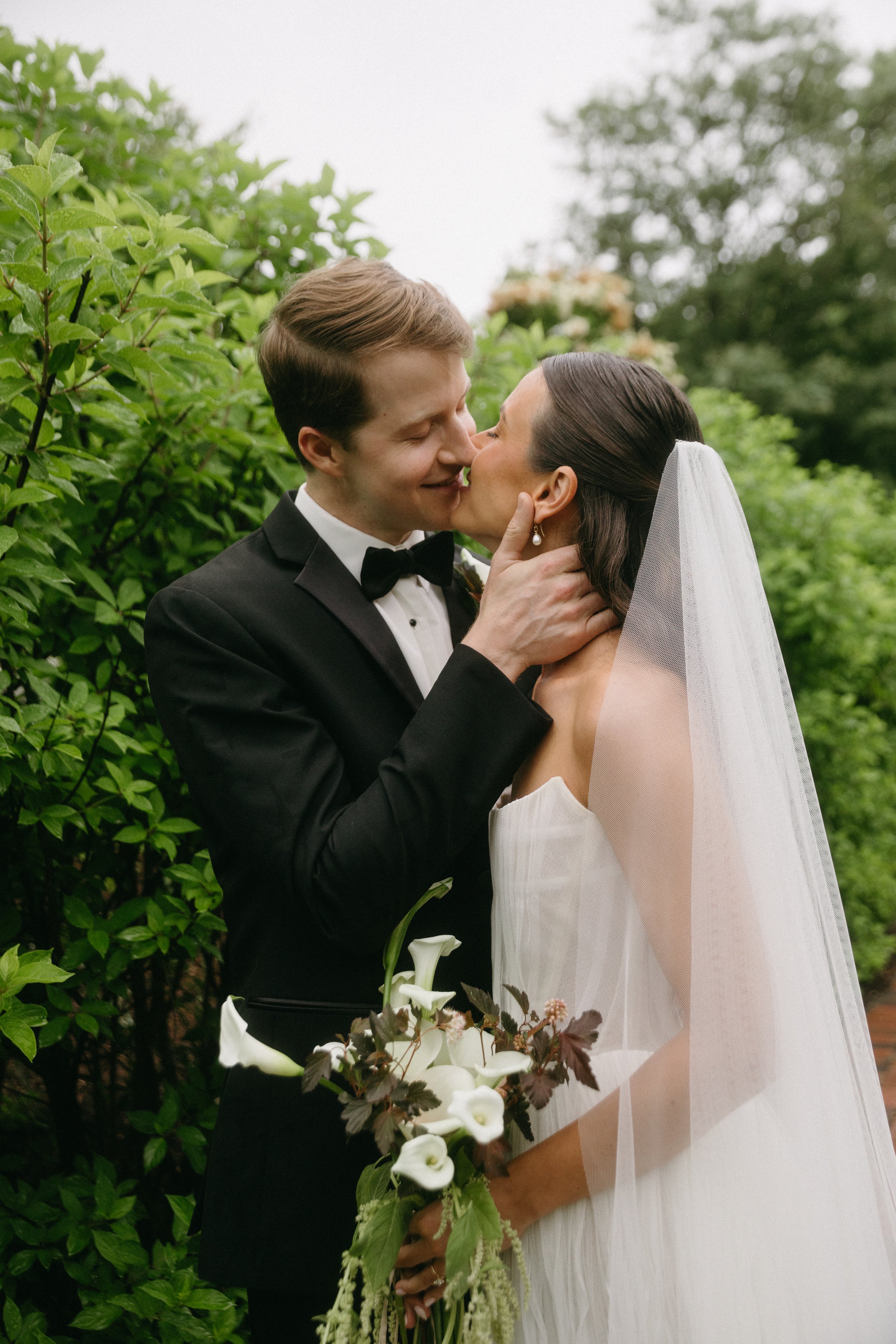 A bride and groom share a kiss outdoors surrounded by green foliage.