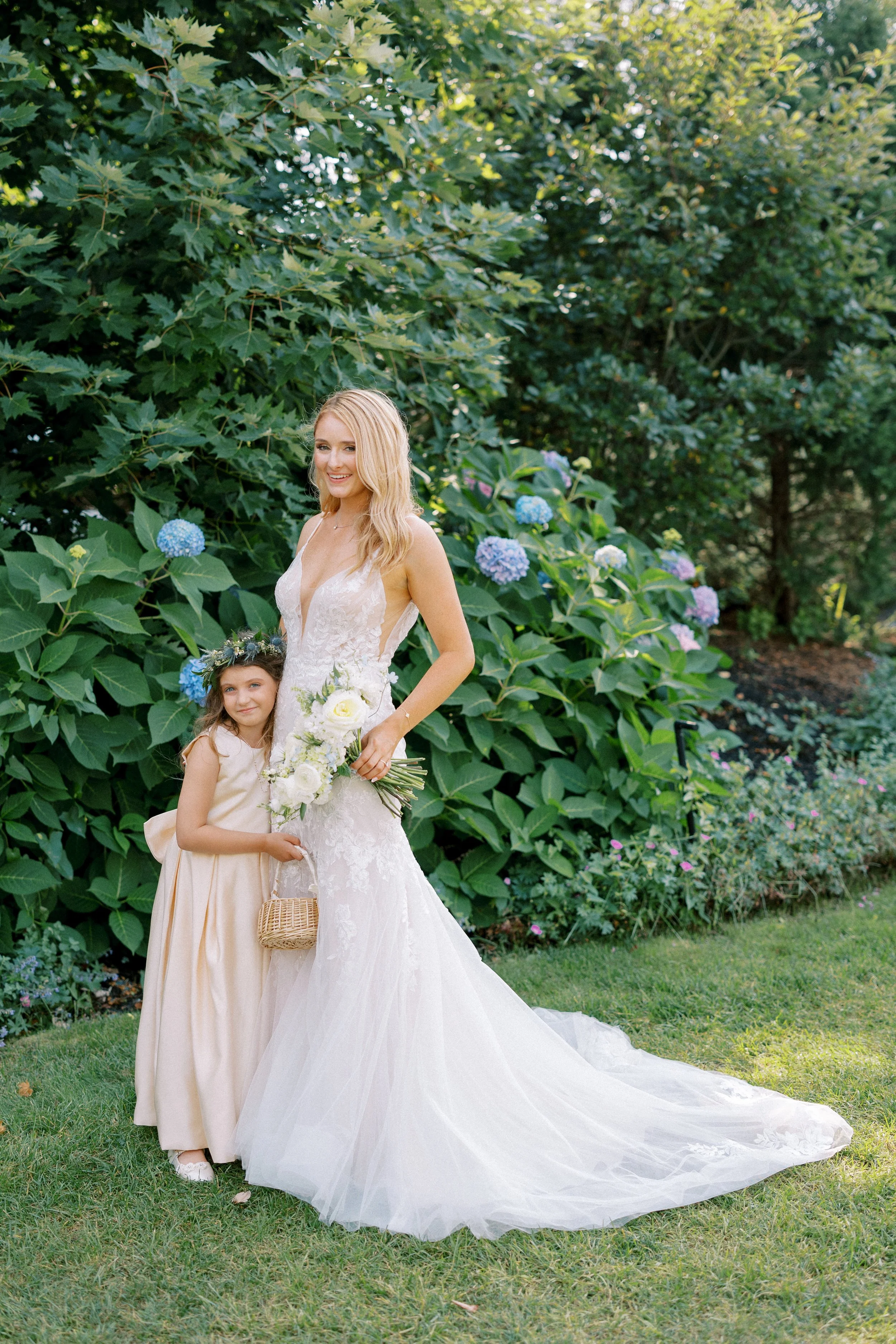 A bride in a white wedding dress holding a bouquet stands next to a young girl in a light-colored dress with a flowers crown in a garden full of greenery and hydrangea flowers.