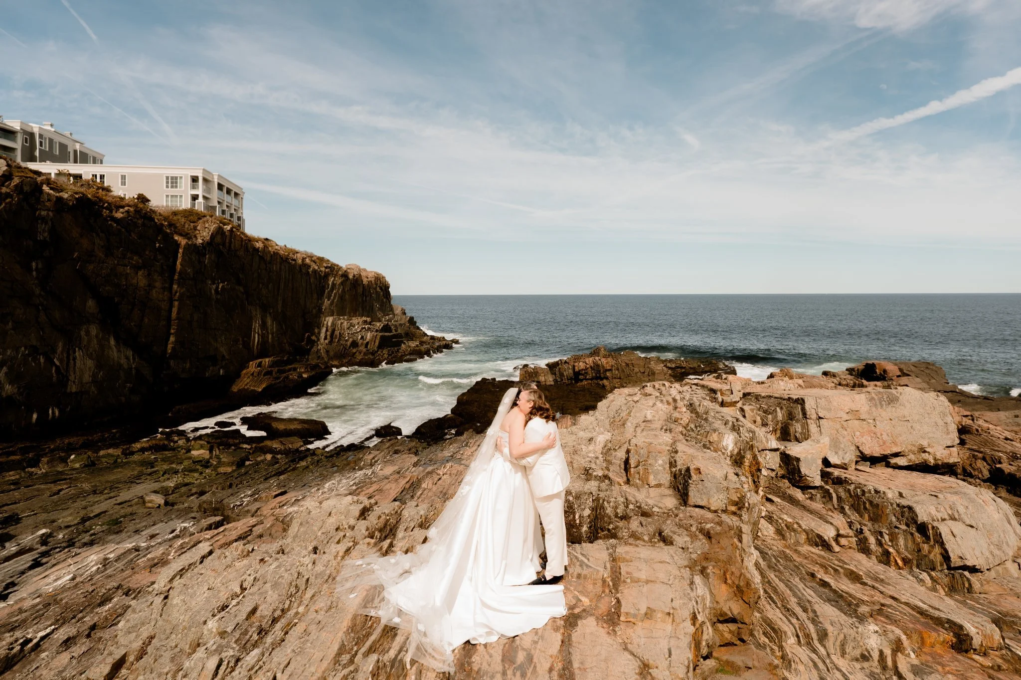 A couple dressed in wedding attire embracing on a rocky shoreline with ocean waves, cliffs, and a partly cloudy sky in the background.