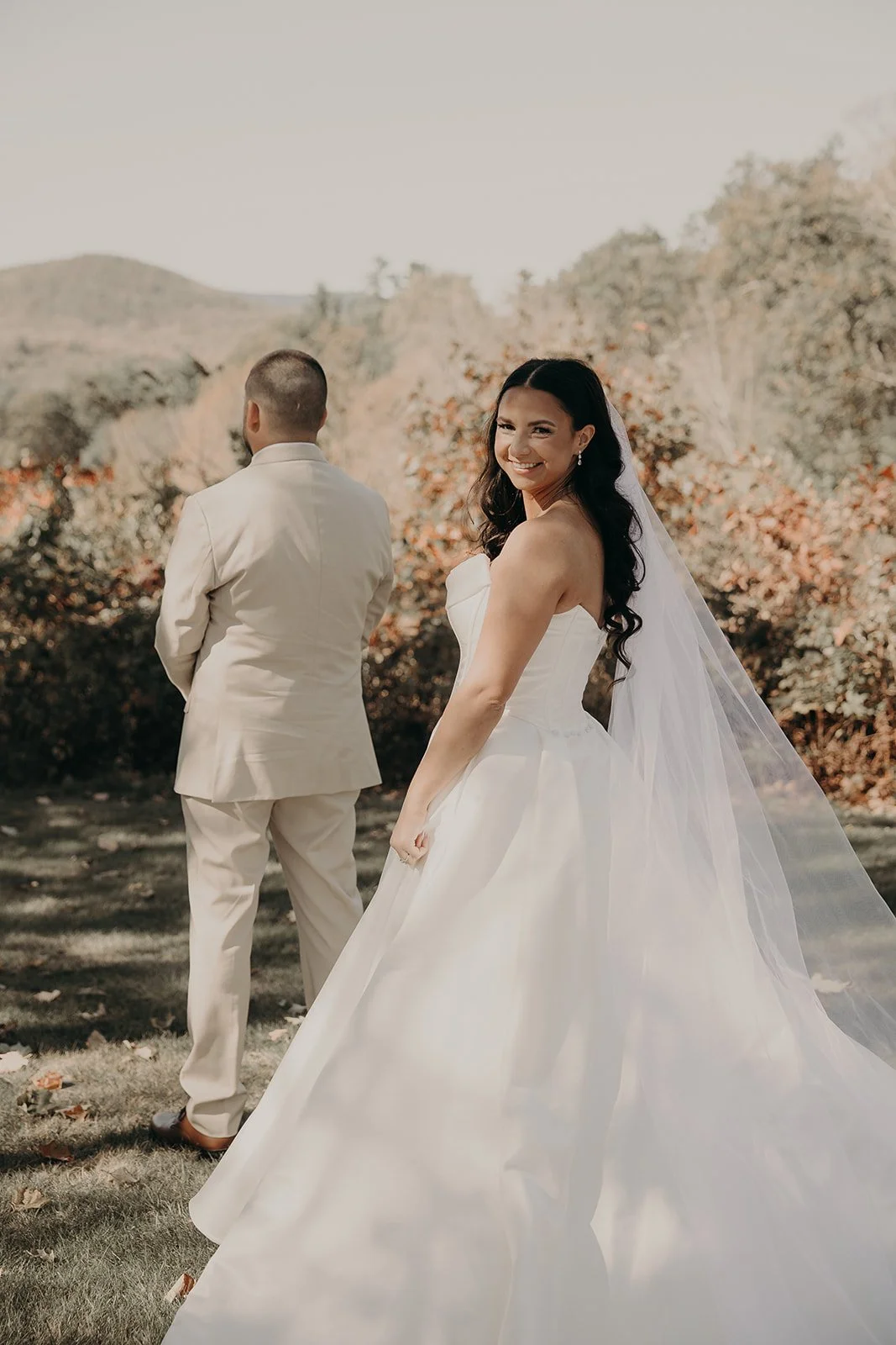 A bride in a white wedding dress smiling and looking back at the camera outdoors, with a groom in a cream suit with his back turned, in a natural setting with trees and mountains in the background.
