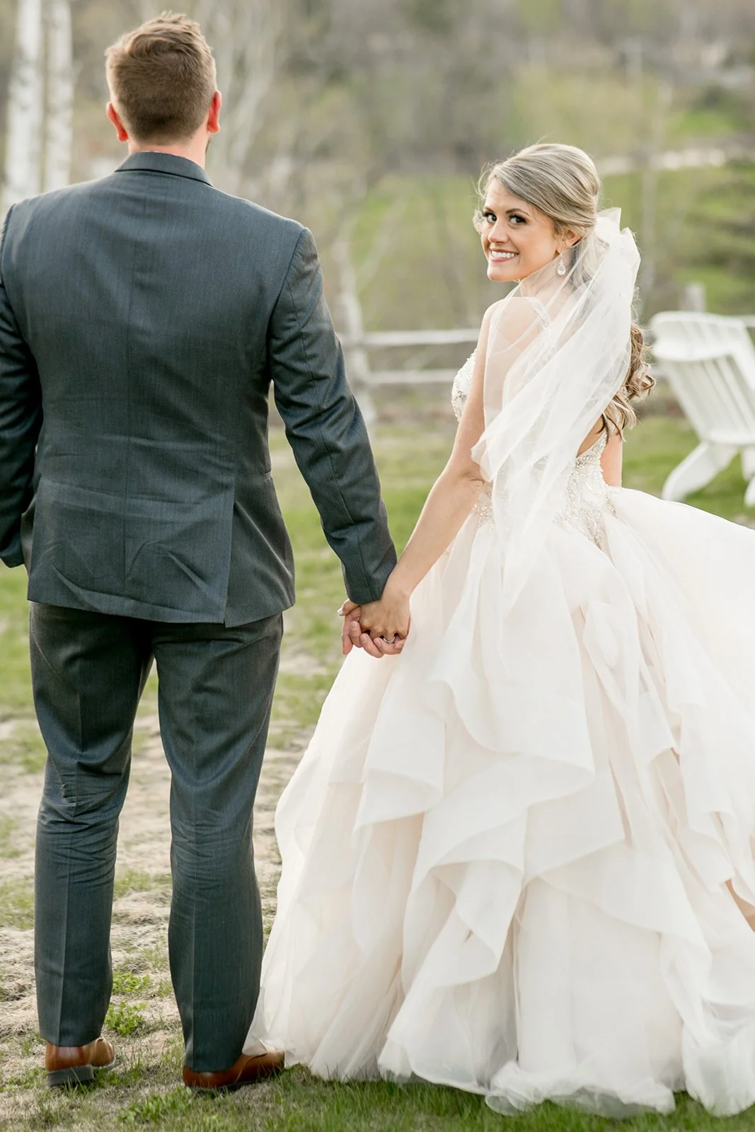 A bride with blonde hair and a veil on is holding her husbands hand is smiling as she looks over her left shoulder back at the camera as he looks straight ahead on their wedding day on May 11, 2019