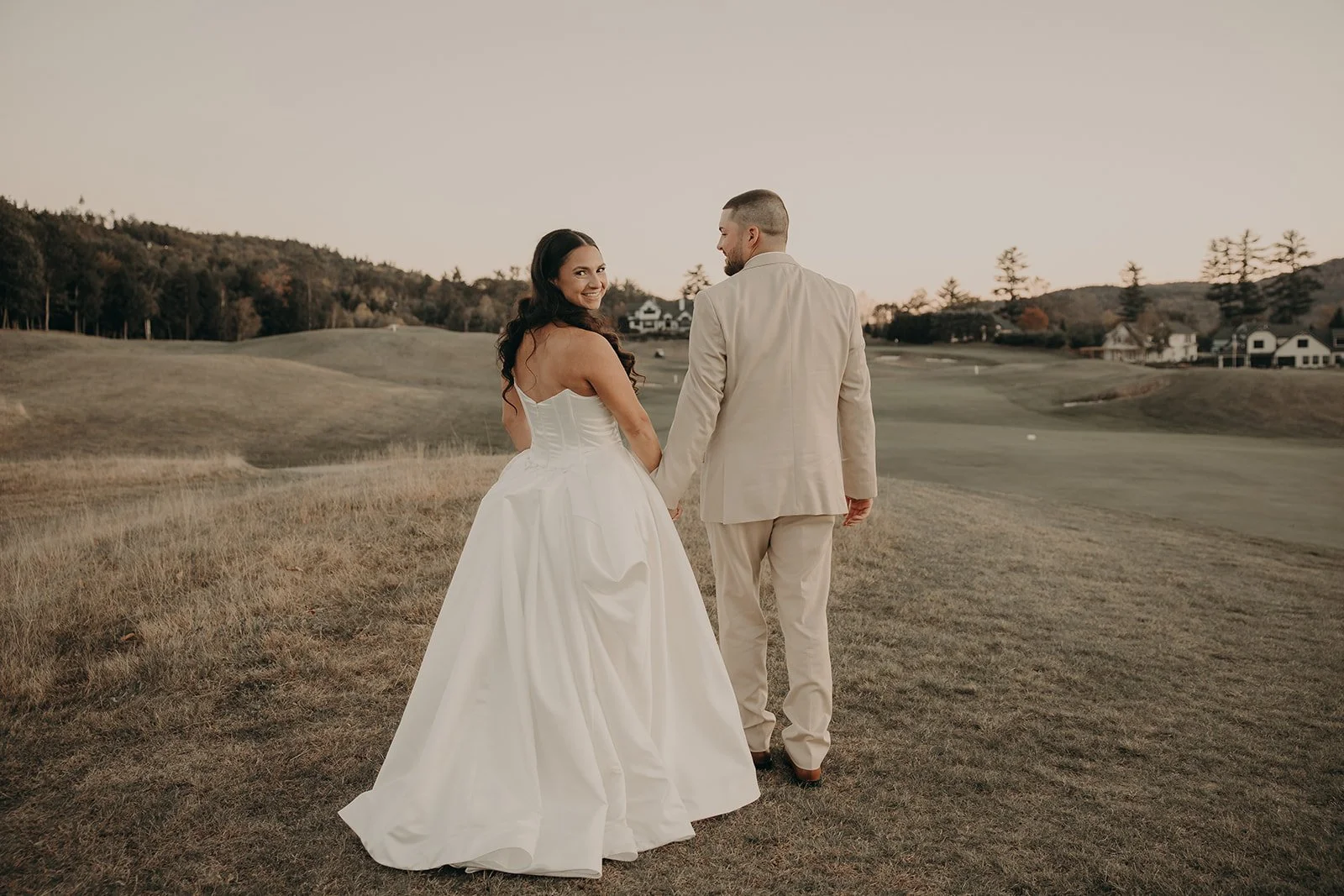 A bride and groom walking hand in hand on a golf course during sunset, dressed in wedding attire, with open landscape, hills, and houses in the background.