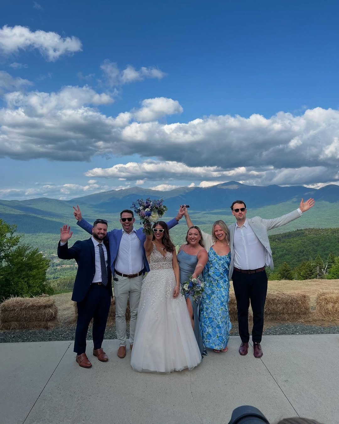 Group of six friends at a wedding, outdoors with mountains and cloudy sky in the background, celebrating with joyful expressions and peace signs.