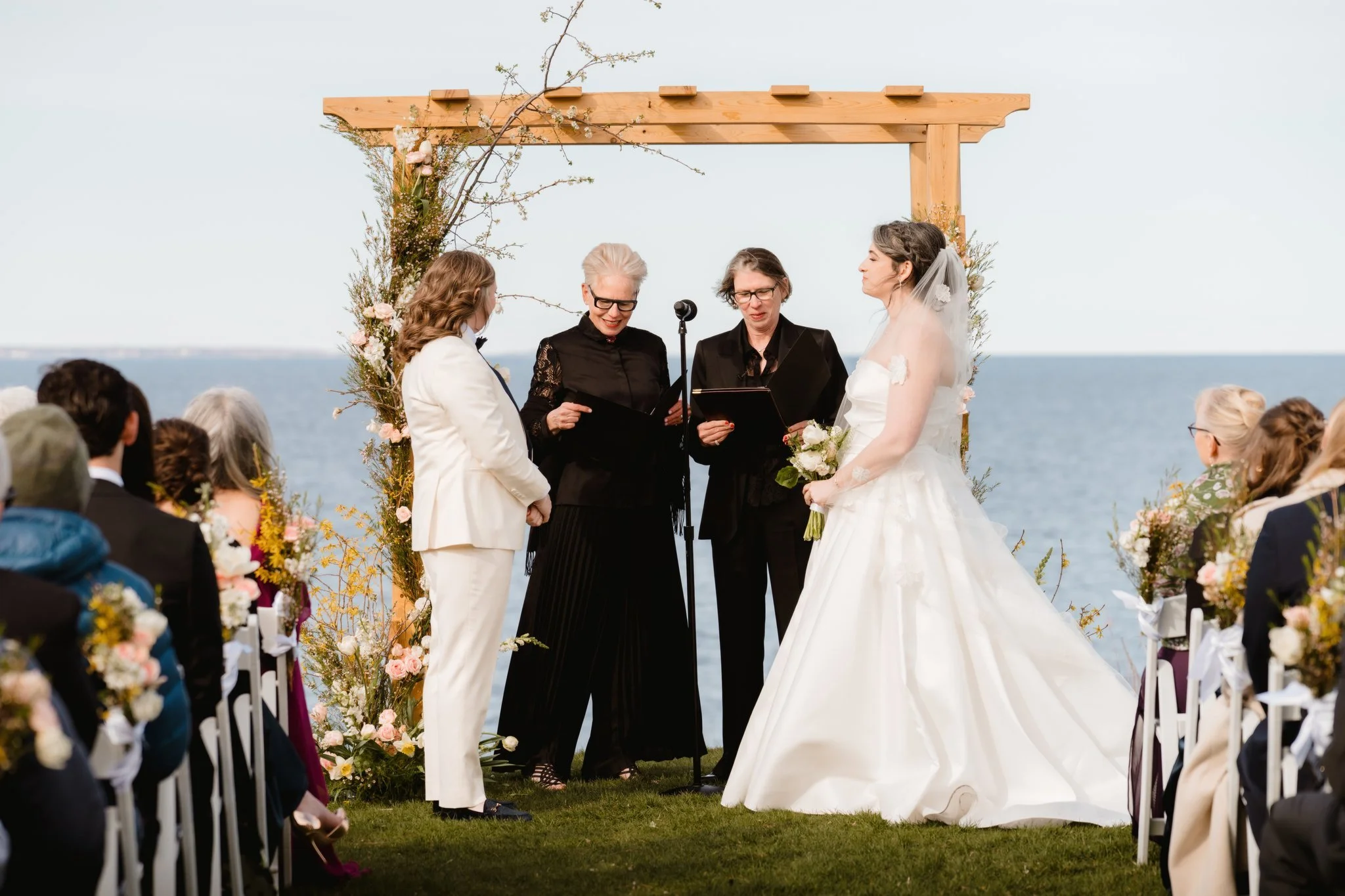 A wedding ceremony taking place outdoors by the water, with two women, one in a white suit and the other in a black outfit, exchanging vows under a wooden arch decorated with flowers. The bride is wearing a white wedding dress and veil, and the officiant is reading from a black folder. Guests are seated on either side of the aisle, and the sky is clear.