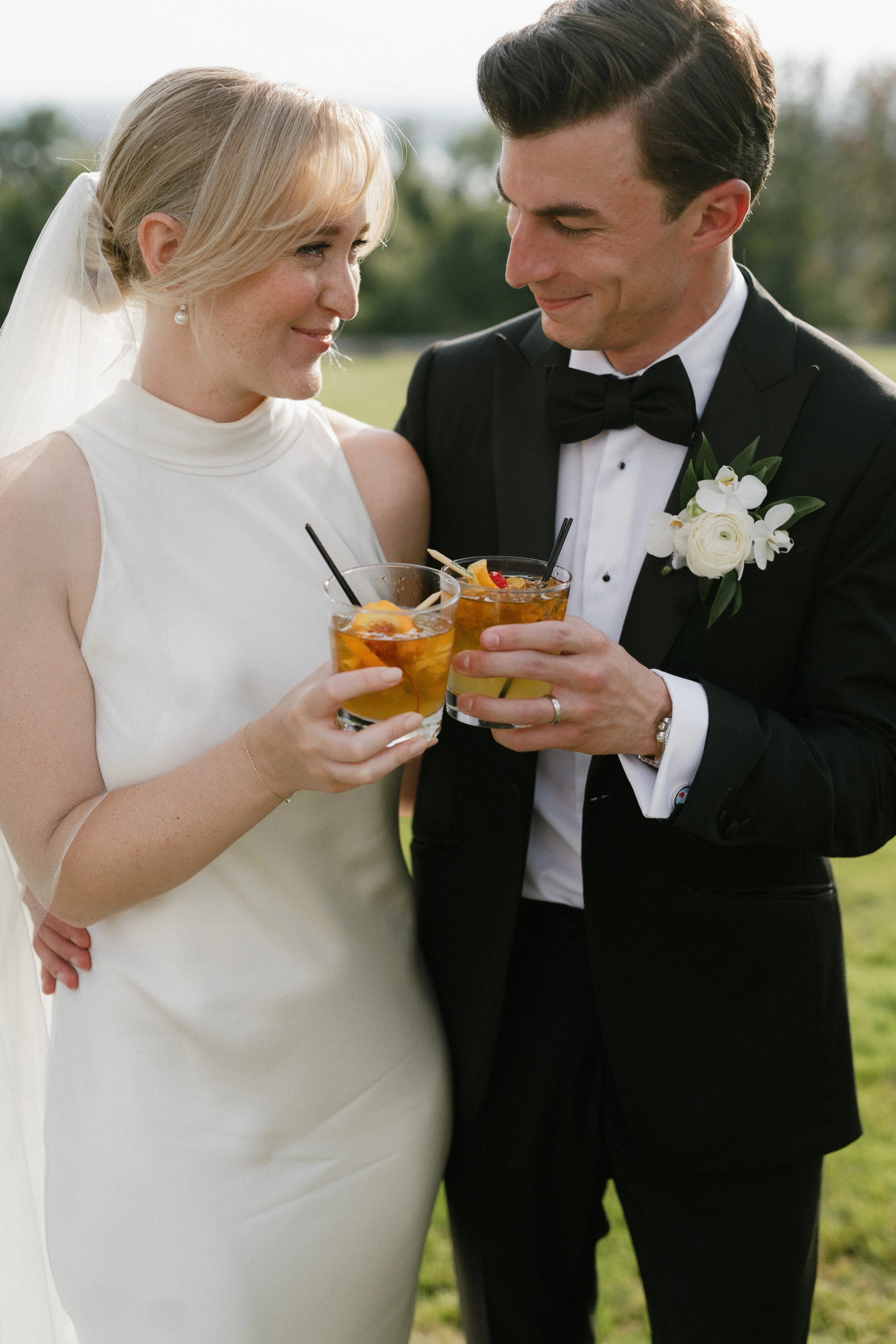 A newlywed couple outdoors, smiling and holding glasses of iced tea, dressed in wedding attire with the bride in a white gown and veil, and the groom in a black tuxedo with a boutonniere, during a celebration or reception.